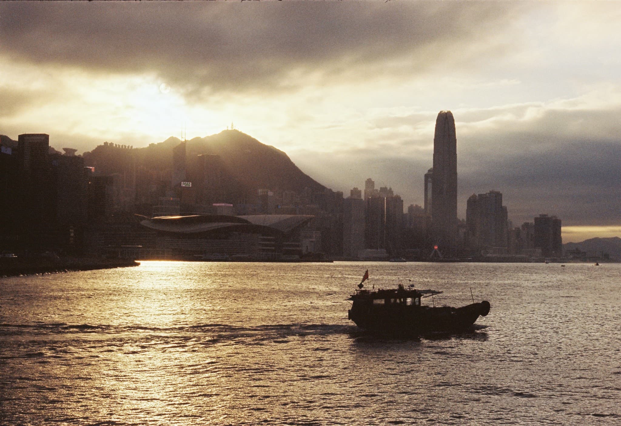 A boat floats on the water in the foreground with a city skyline featuring a tall skyscraper and a mountain illuminated by the setting sun in the background.