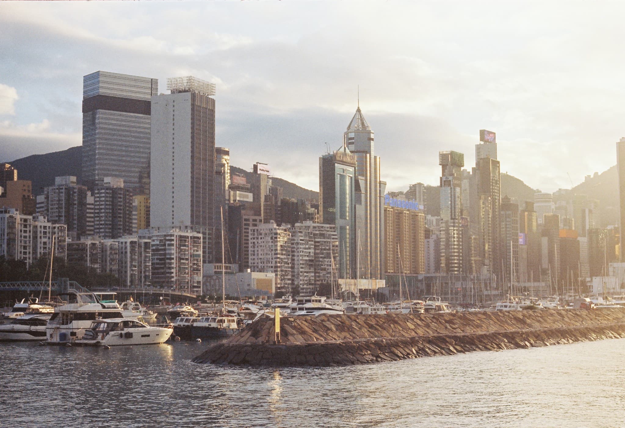 A harbor filled with docked boats and a stone jetty sits in front of a dense urban skyline of high-rise buildings and mountains under a cloudy sky.