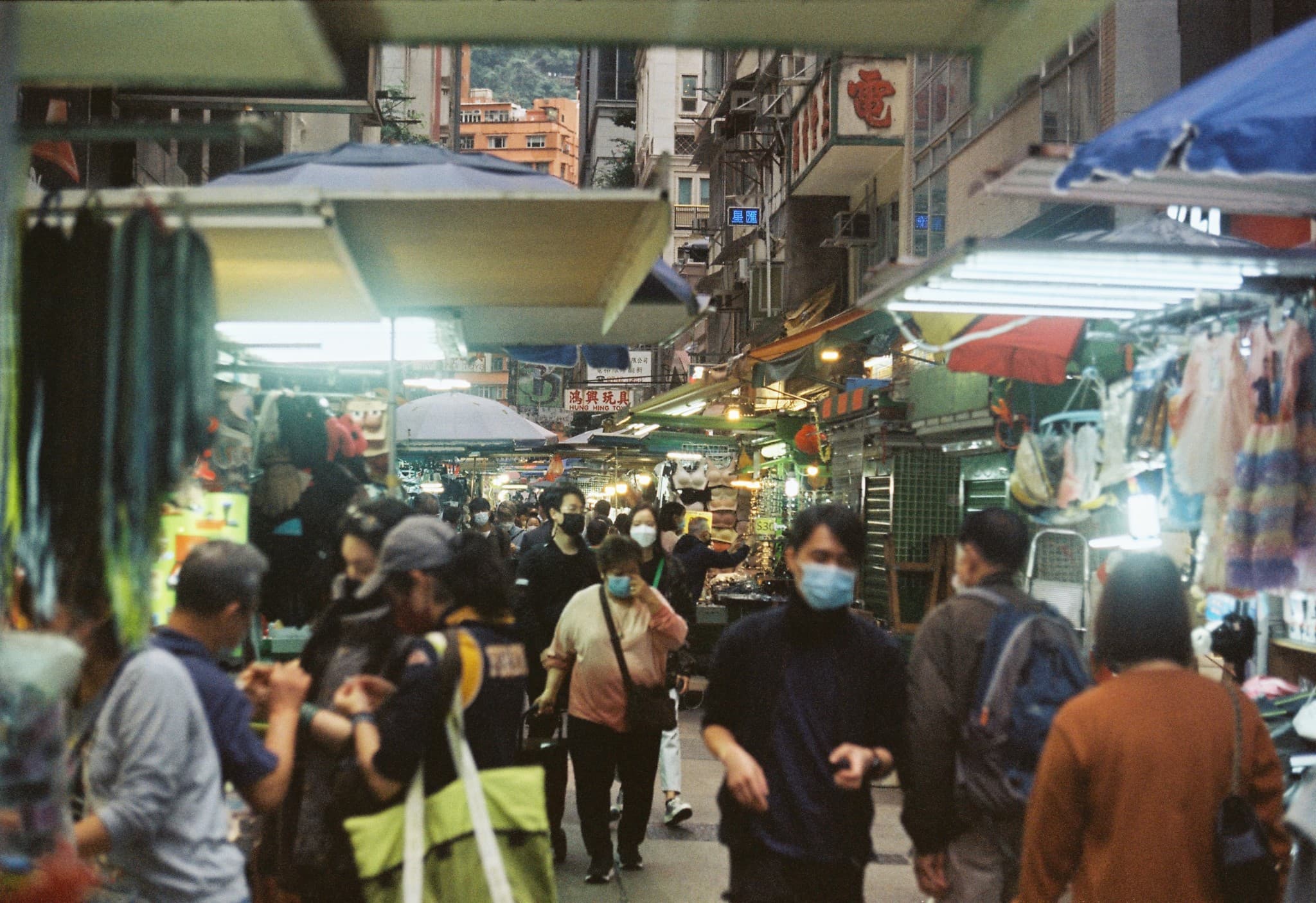 A crowded, covered street market aisle filled with people walking between stalls selling clothing and other goods.