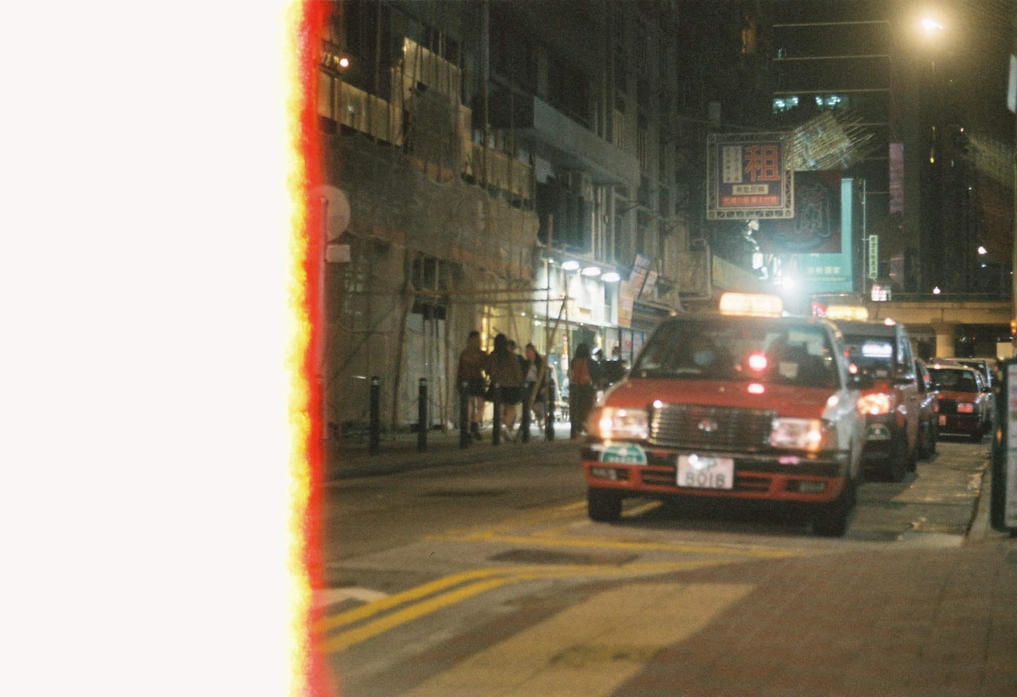 A red taxi drives down a city street at night past pedestrians walking on a sidewalk near a building with scaffolding.
