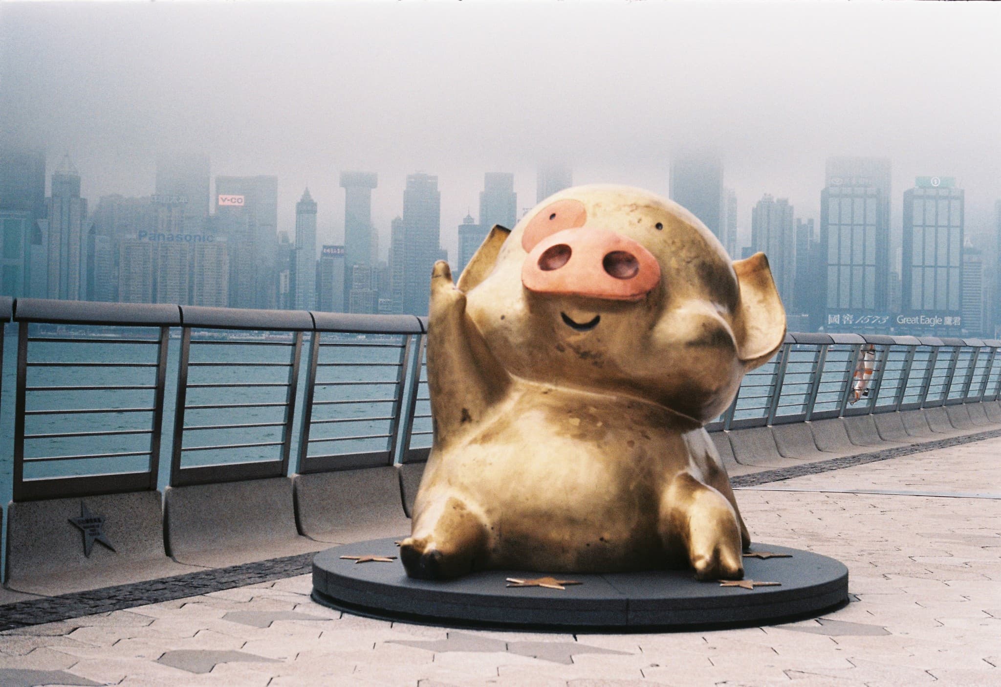 A golden statue of a smiling pig sits on a circular base surrounded by stars, positioned on a waterfront walkway with a foggy city skyline in the background.
