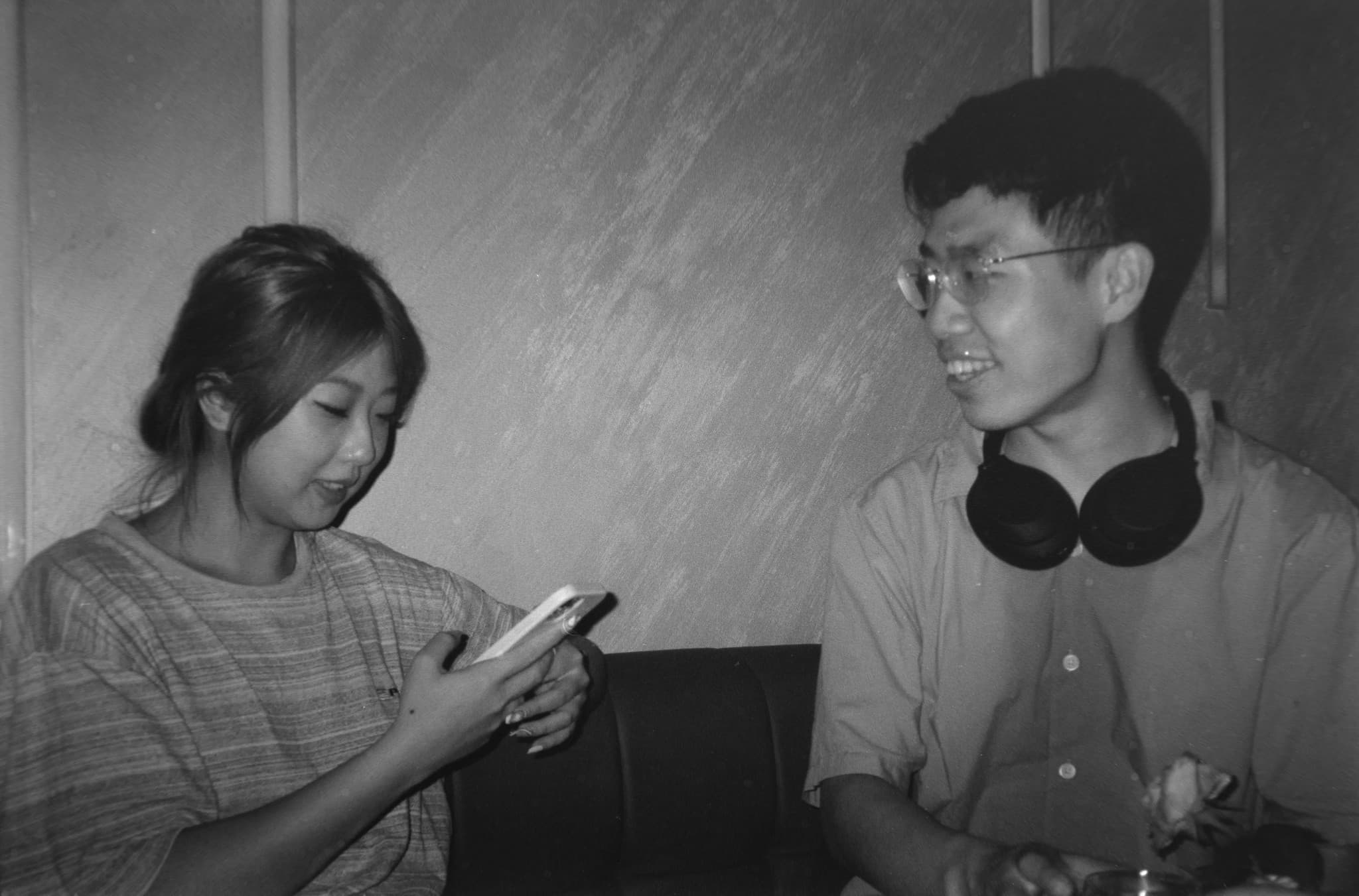 A black and white photo showing a woman looking at her phone while a smiling man wearing glasses and headphones around his neck looks on.