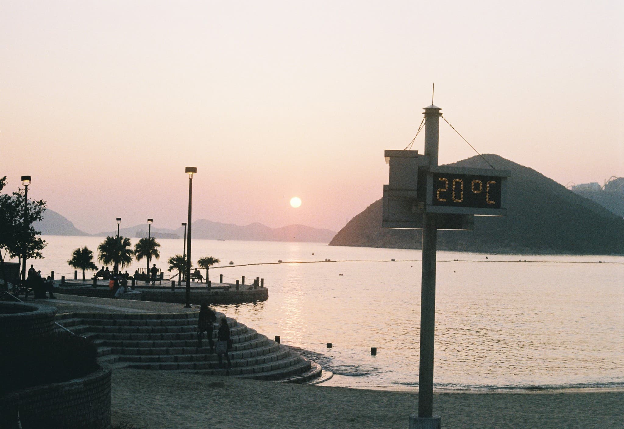 A digital temperature display showing 20°C stands in the foreground of a beach at sunset, overlooking calm water and silhouetted hills.