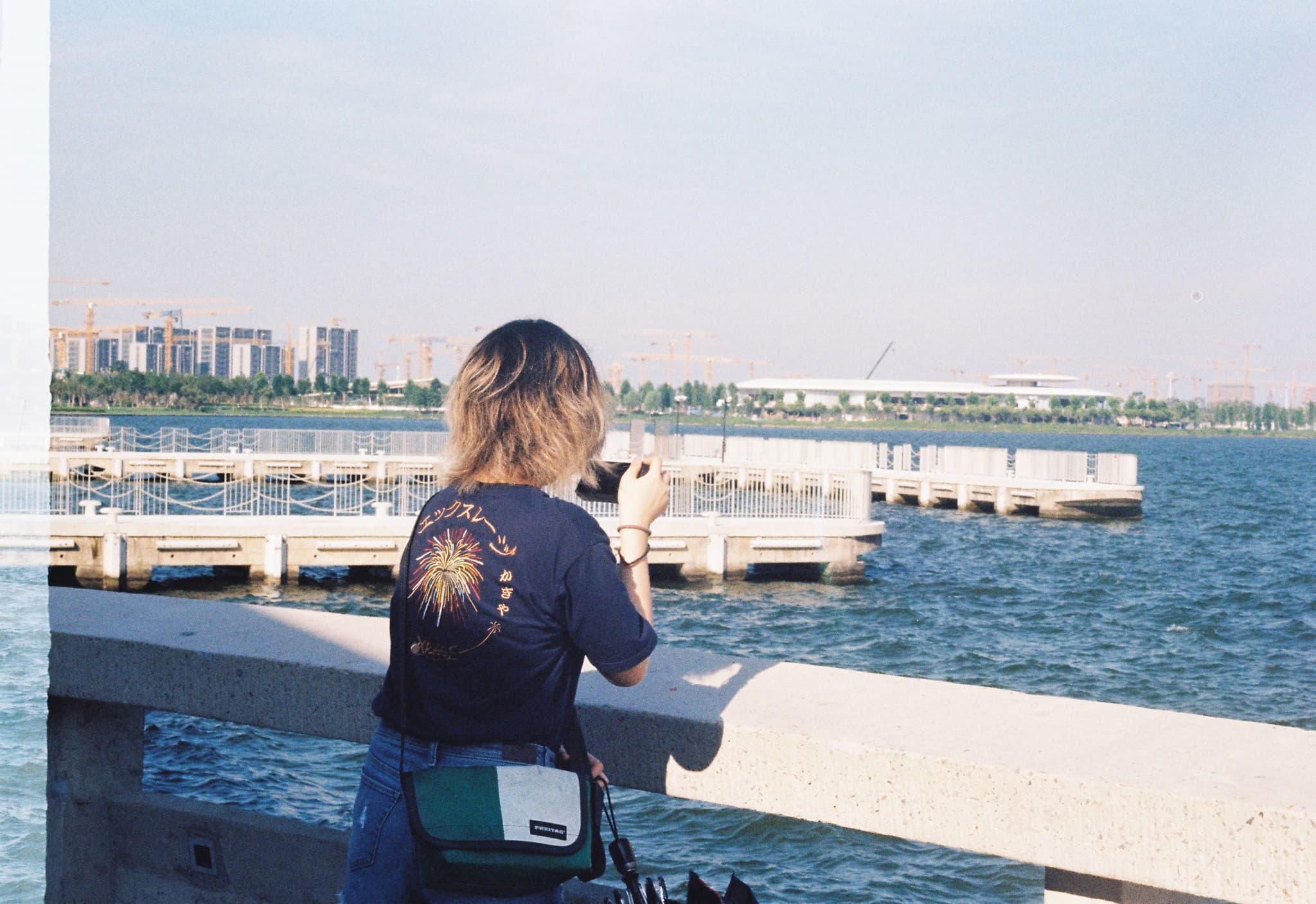 A person with blonde hair stands on a bridge overlooking a body of water with a pier and a city skyline featuring construction cranes in the distance.