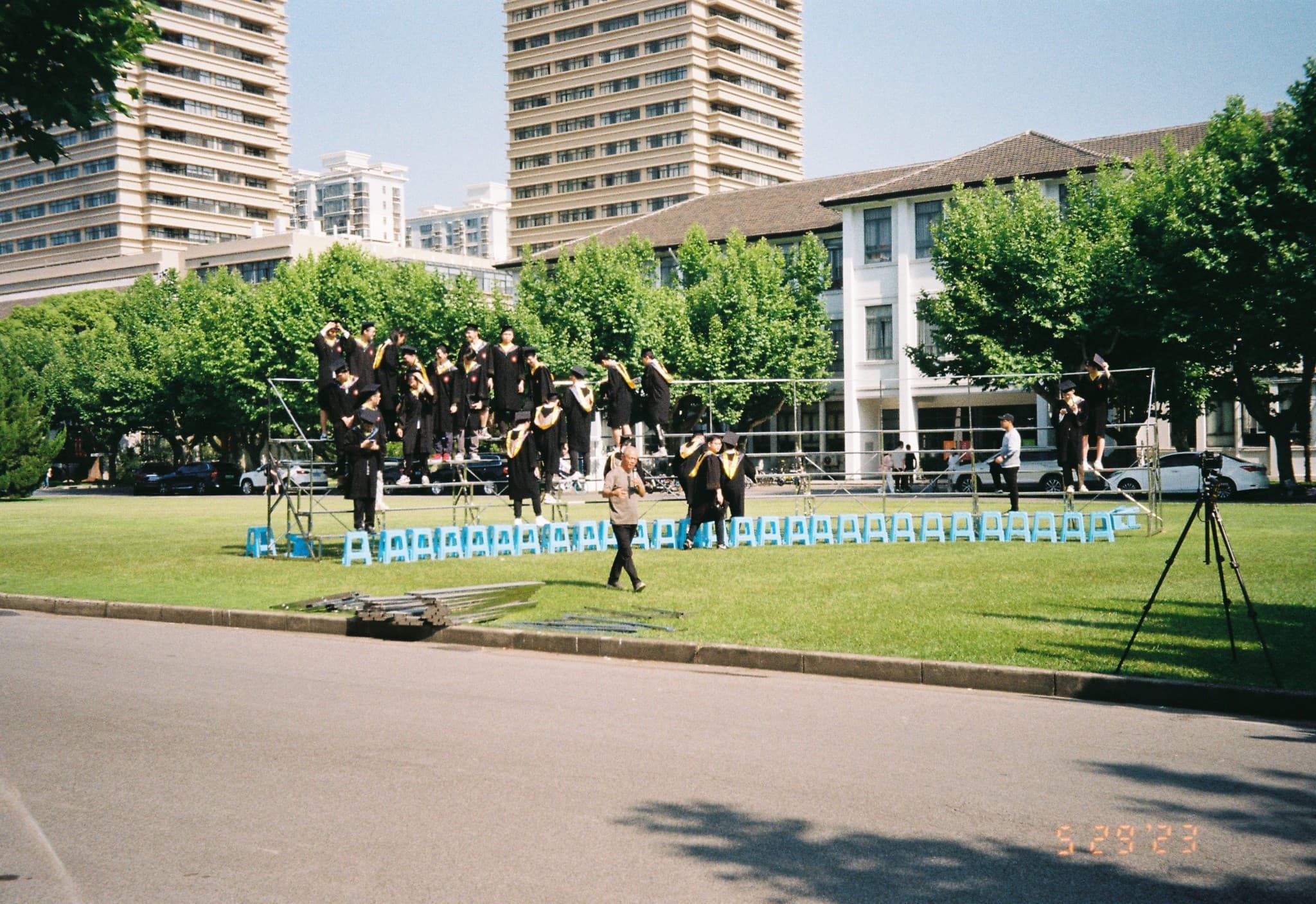 Graduates in black gowns pose on a metal scaffold structure on a grassy lawn, with a row of blue plastic chairs lined up at the base.