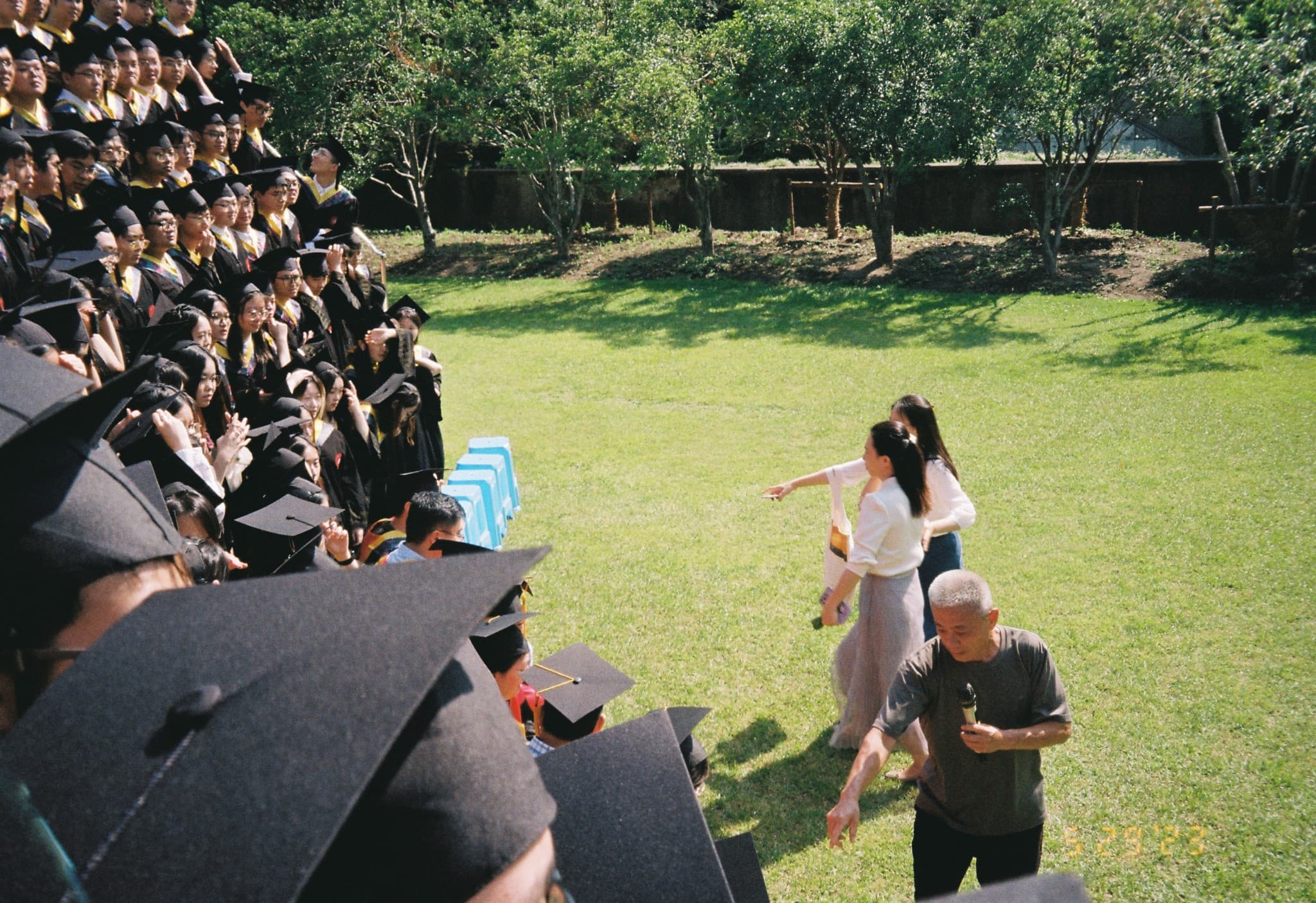 A large group of graduates in caps and gowns sits on bleachers on the left, while a man holding a microphone and two women stand on a grassy field to the right.