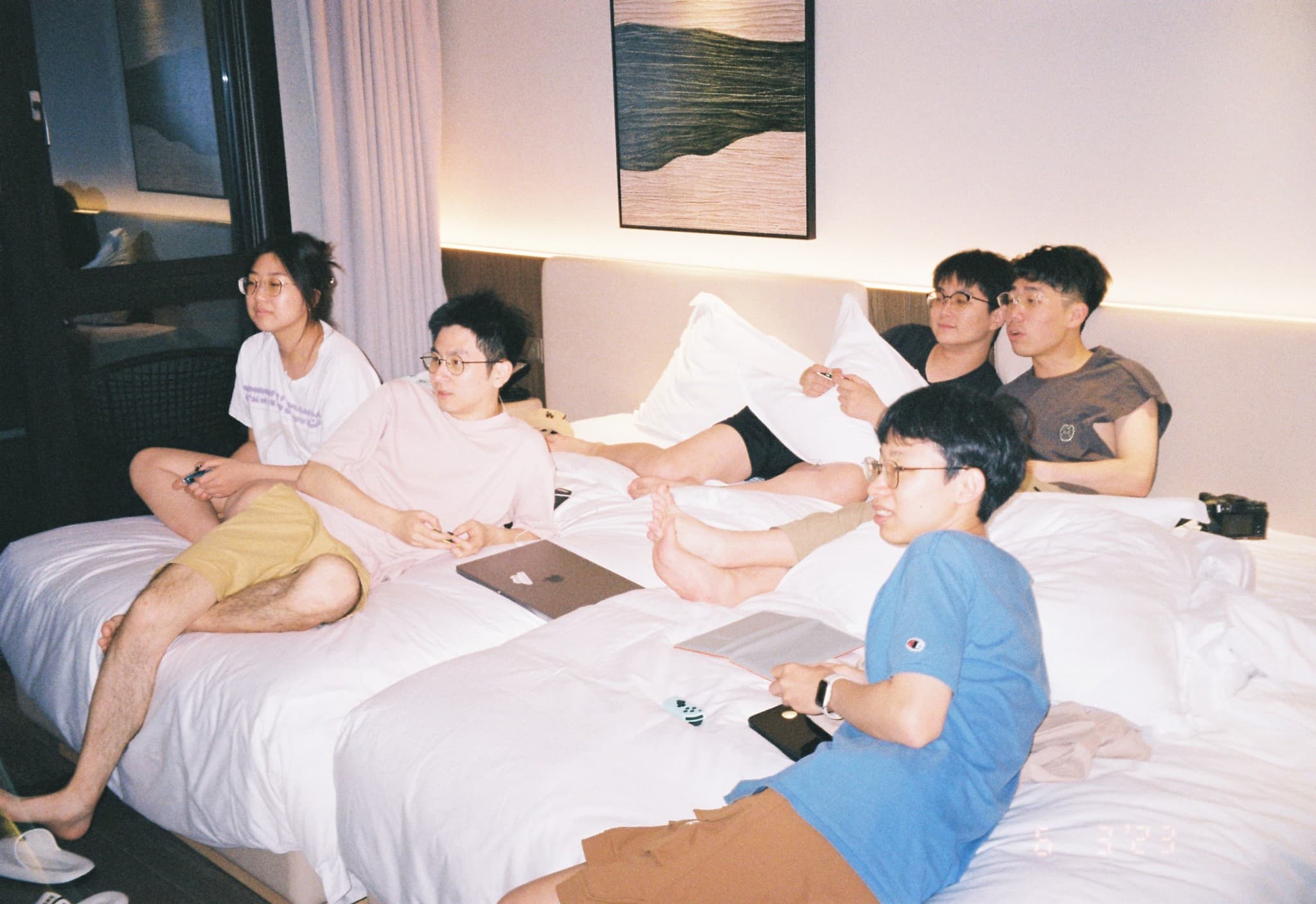 Five young adults relax on beds in a hotel room, watching something off-camera while holding remote controls.