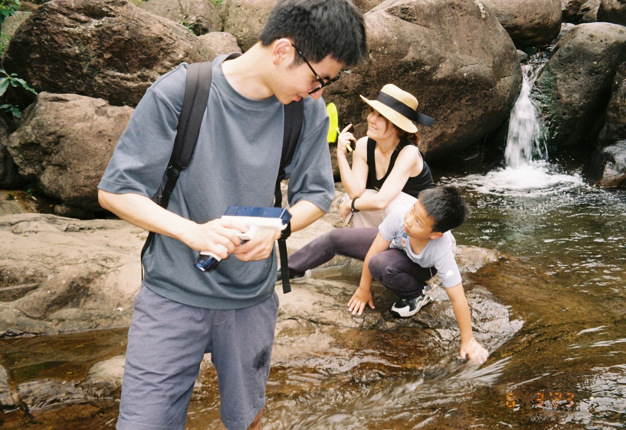 A young man holding a white toy water gun stands next to a woman in a hat and a young boy crouching near a rocky stream.