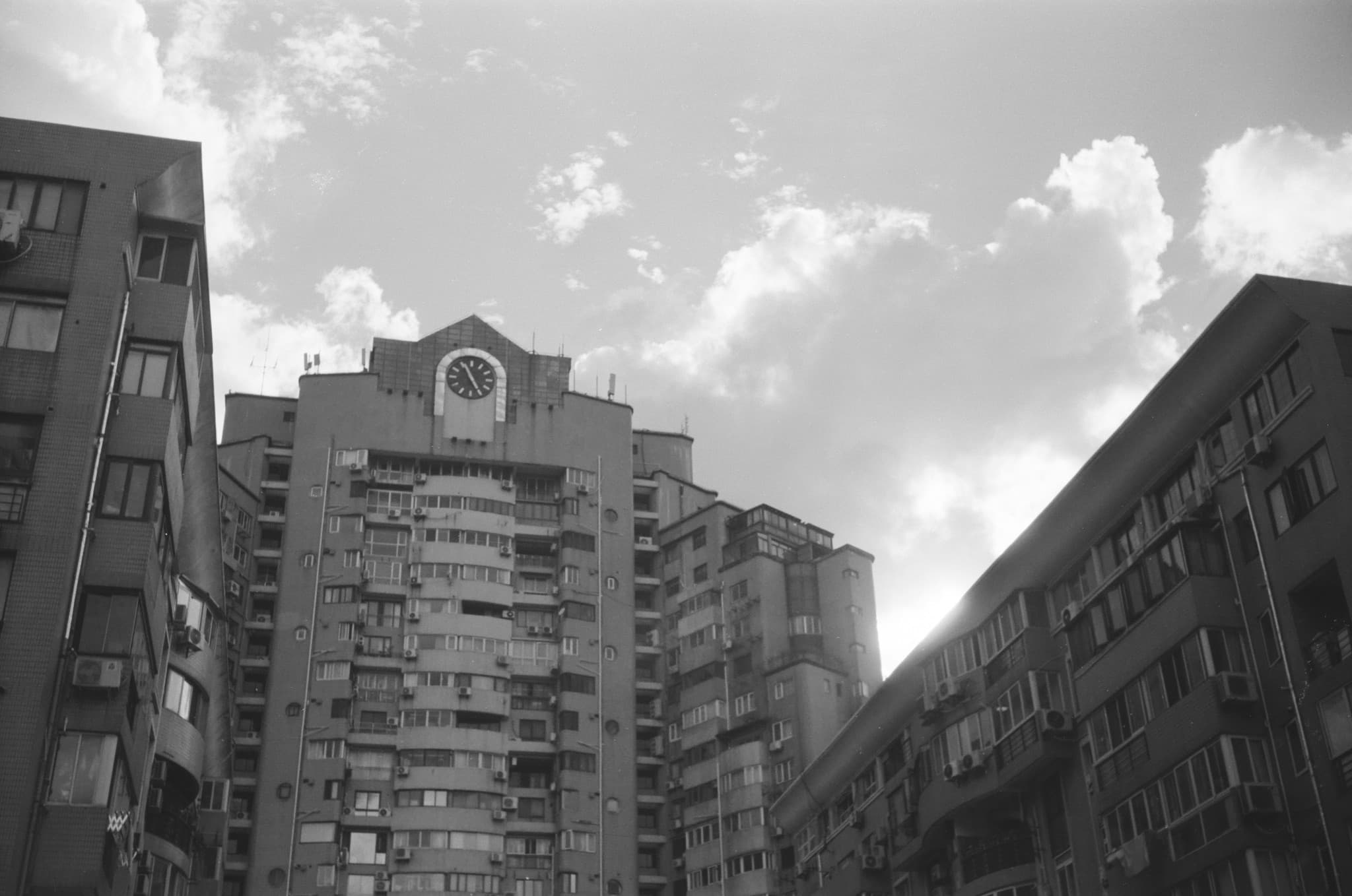 A low-angle black and white photograph showing three multi-story apartment buildings against a cloudy sky, with a clock face near the top of the central building.