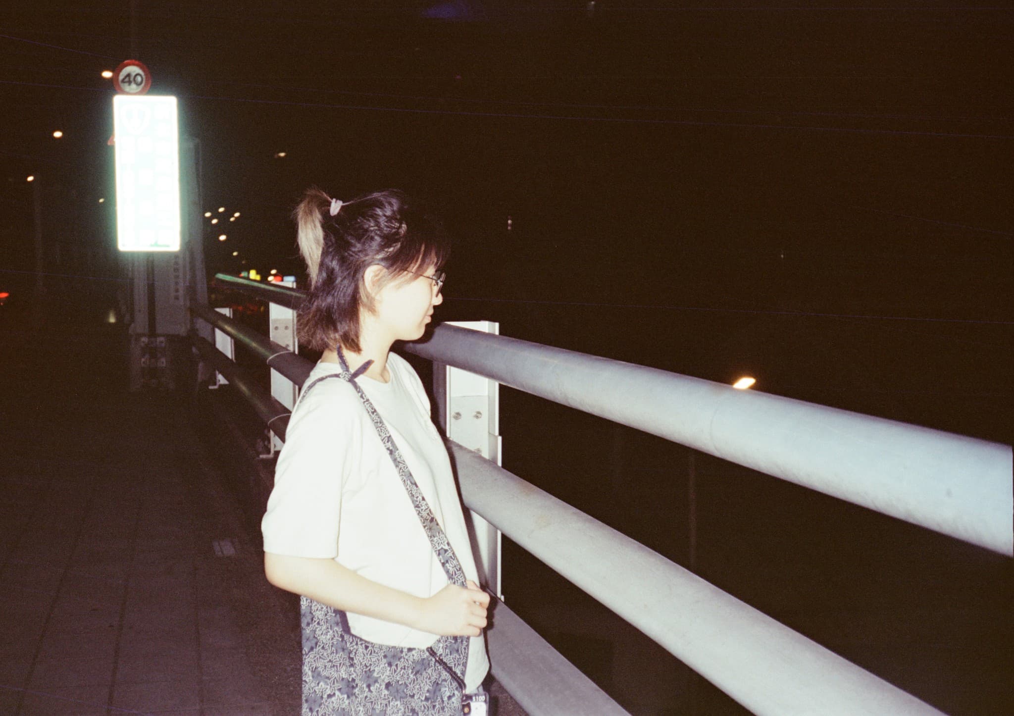 A young woman with glasses and a ponytail stands in profile next to a metal railing on a bridge at night.