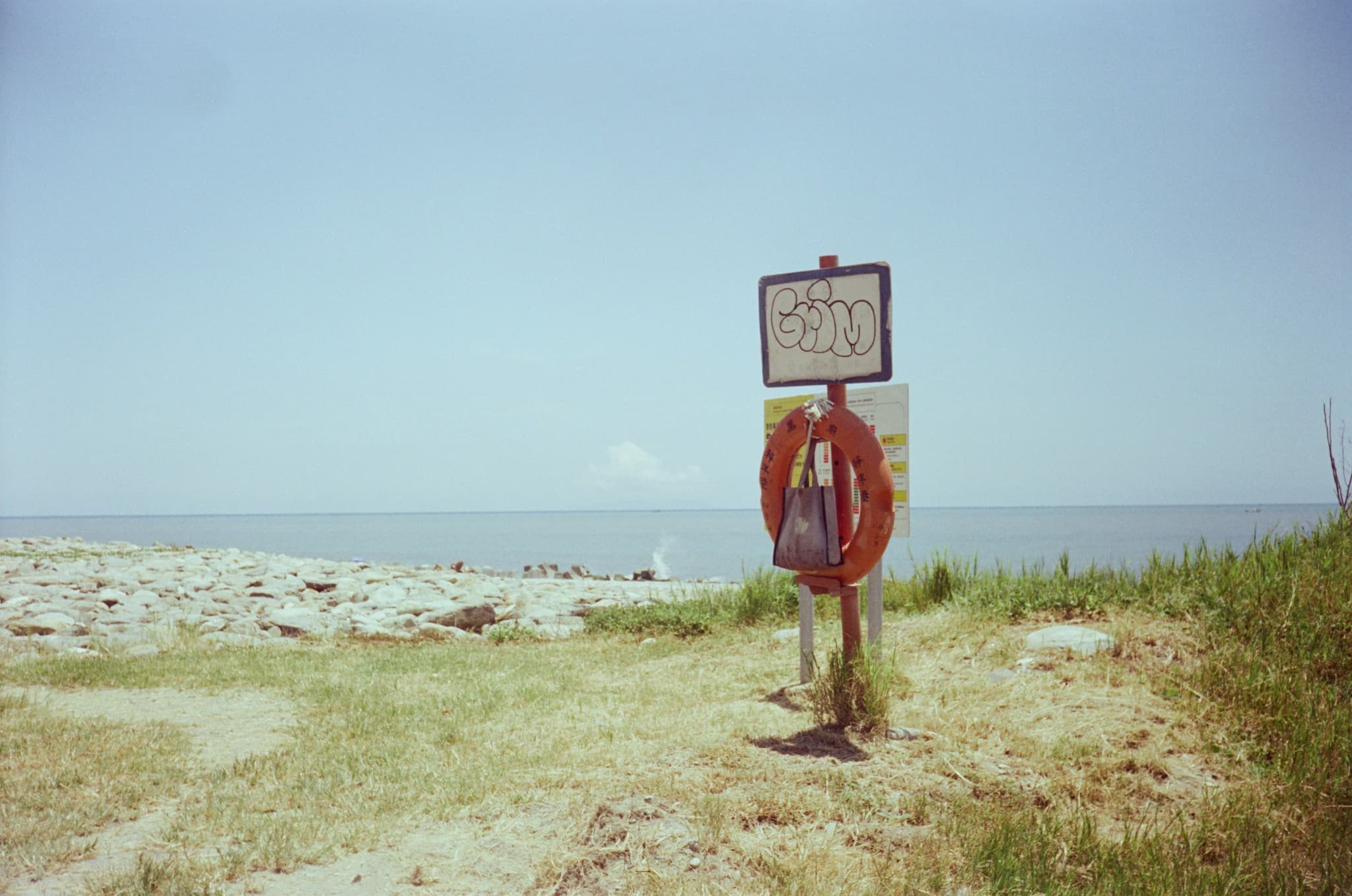 A red lifebuoy hangs from a signpost on a grassy verge, with a rocky beach and calm ocean in the background.