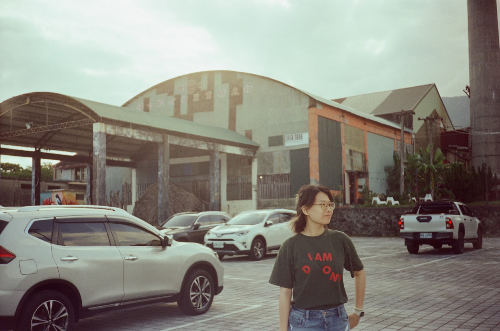 A young woman wearing glasses stands in a paved lot with several parked cars, set against a backdrop of a large, weathered industrial building and a tall chimney.