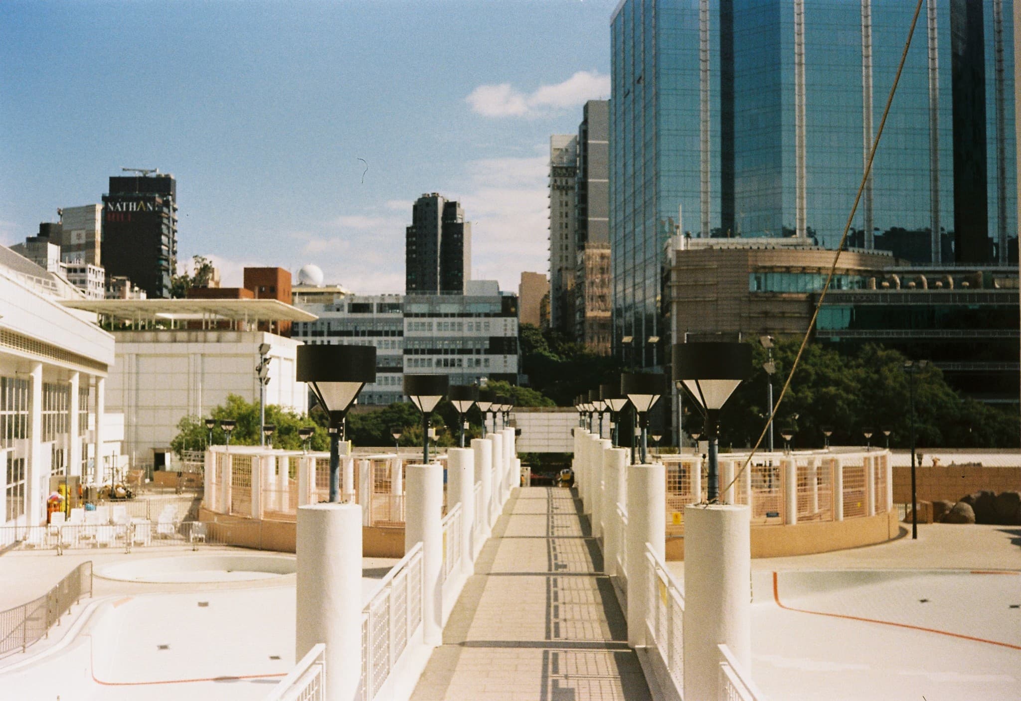 A paved walkway lined with white pillars and black lamps leads toward a city skyline featuring a large blue glass skyscraper on the right and other urban buildings in the distance.