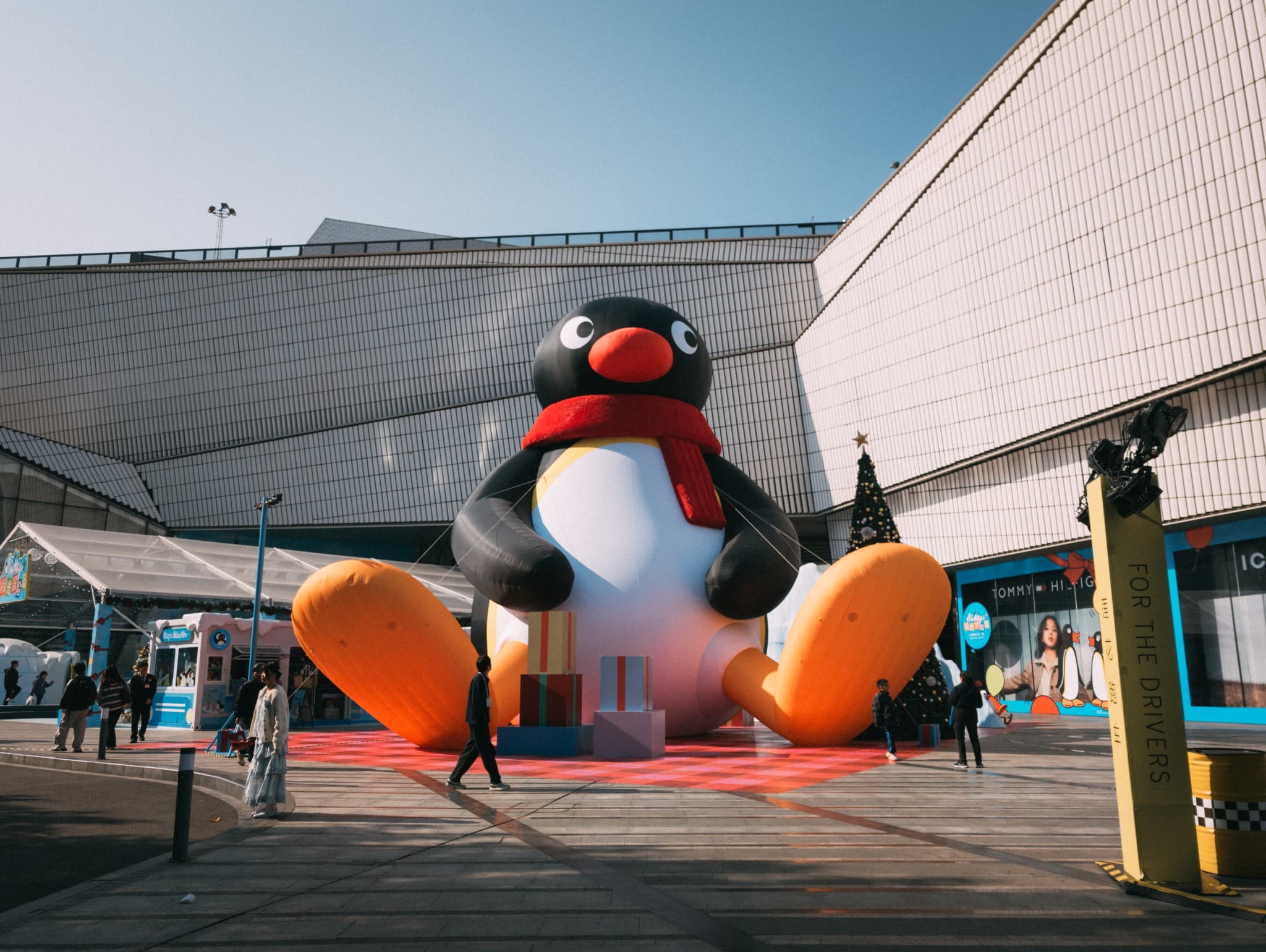 A giant inflatable penguin wearing a red scarf sits on a red rug in an outdoor plaza in front of a shopping mall.