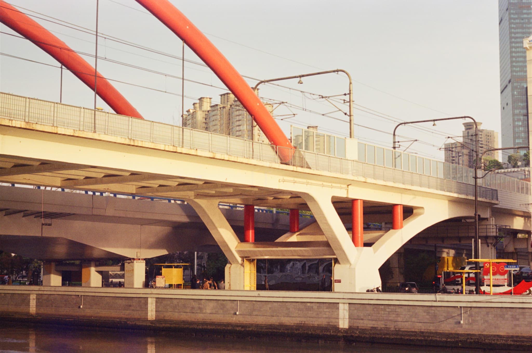 A view looking up at a bridge with large red curved supports and overhead wires, situated above a river with city buildings in the background.