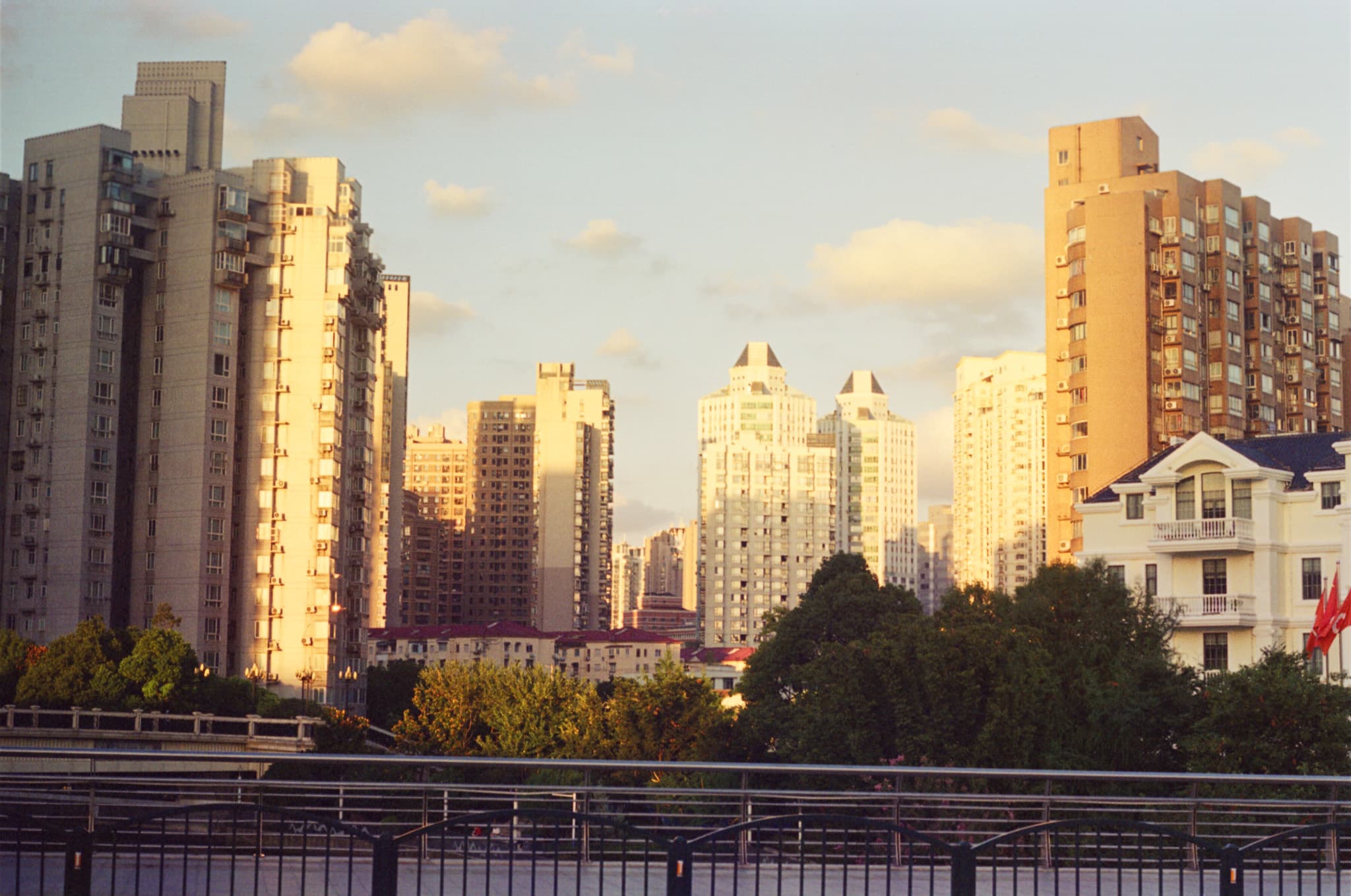 A row of tall apartment buildings is illuminated by warm sunlight, viewed from behind trees and a metal railing.
