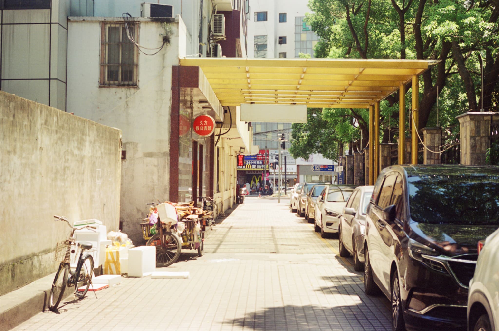 A paved sidewalk stretches down a narrow urban street lined with parked cars on the right and loaded bicycles near a wall on the left, all shaded by a large yellow metal canopy.