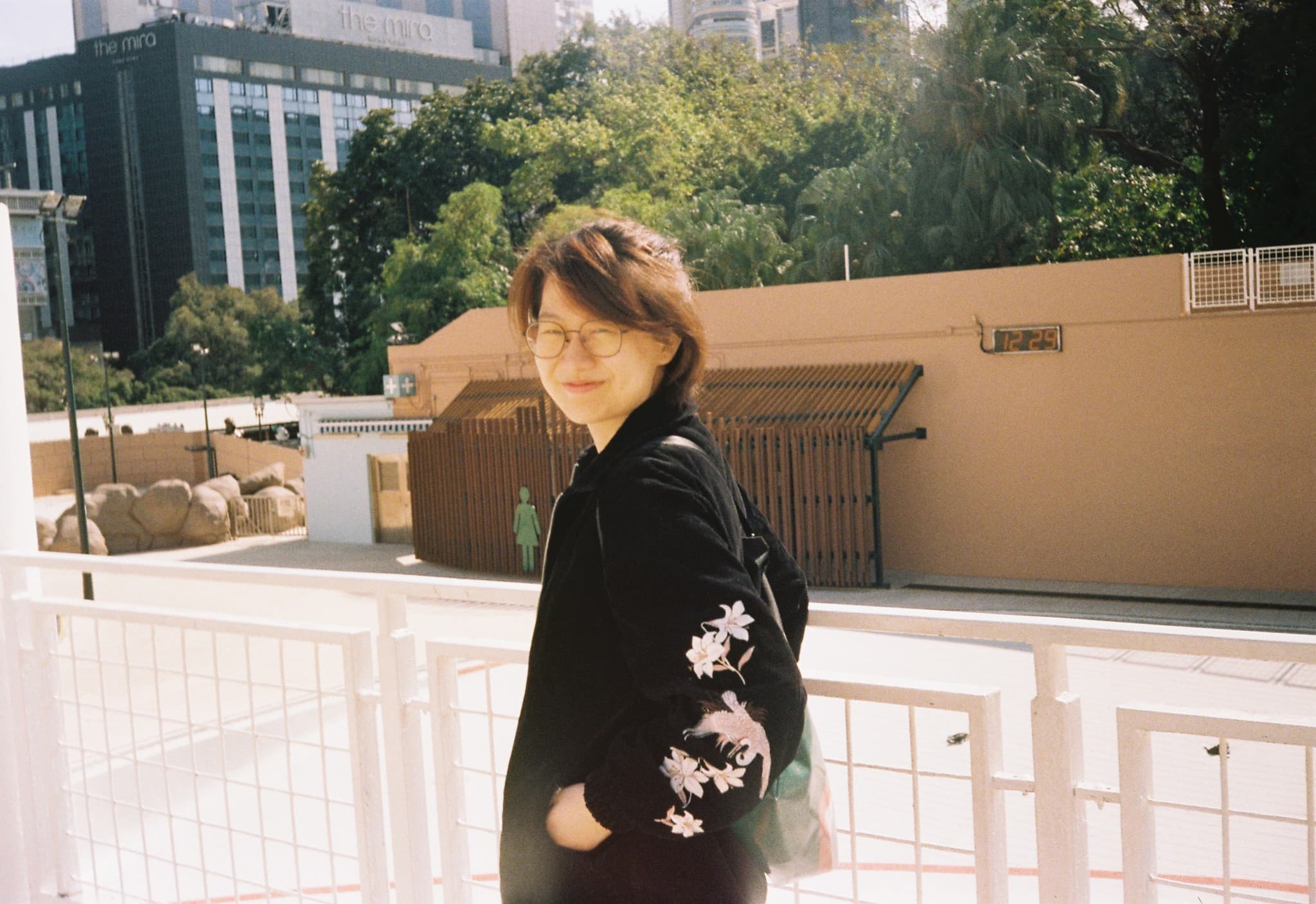 A person with short hair and glasses wearing a black jacket with white embroidery looks back over their shoulder while standing on a sunny outdoor walkway with a city building and trees in the background.