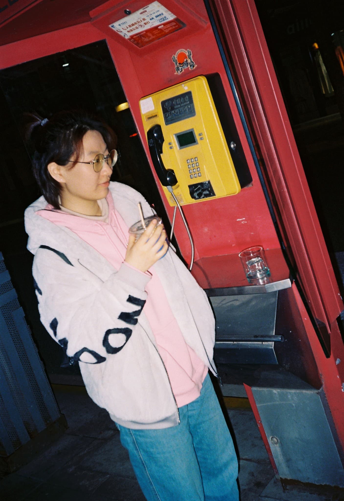 A person wearing a white hoodie and glasses leans over a red phone booth holding a drink next to a yellow payphone.