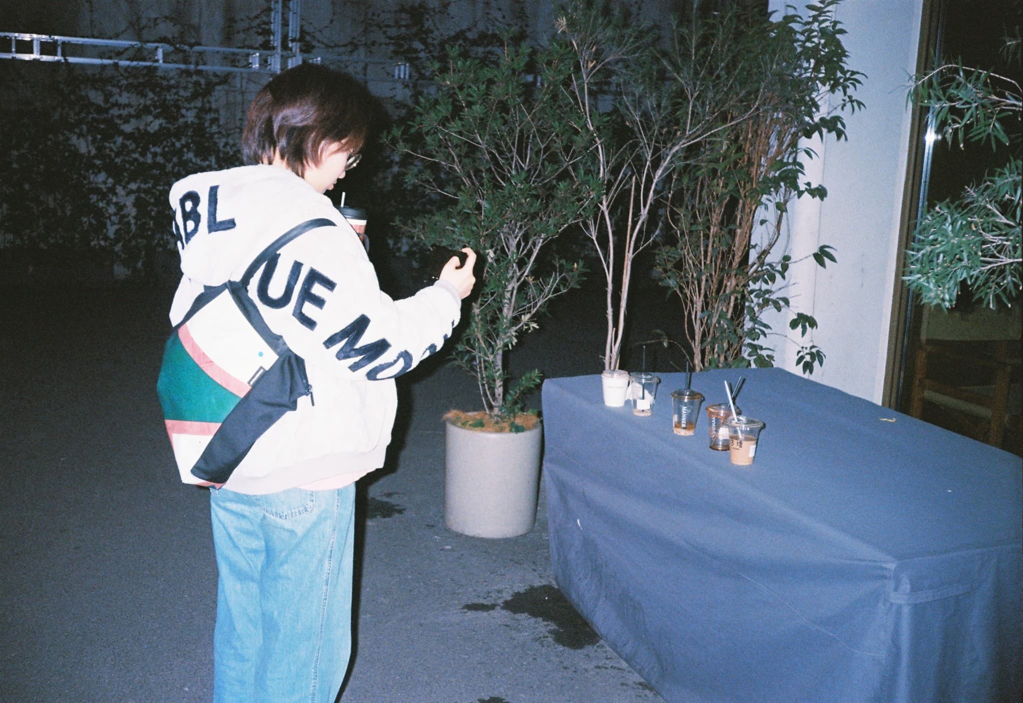 A person in a white hoodie stands next to a long table covered with a blue cloth that holds several plastic cups, looking towards a potted plant at night.