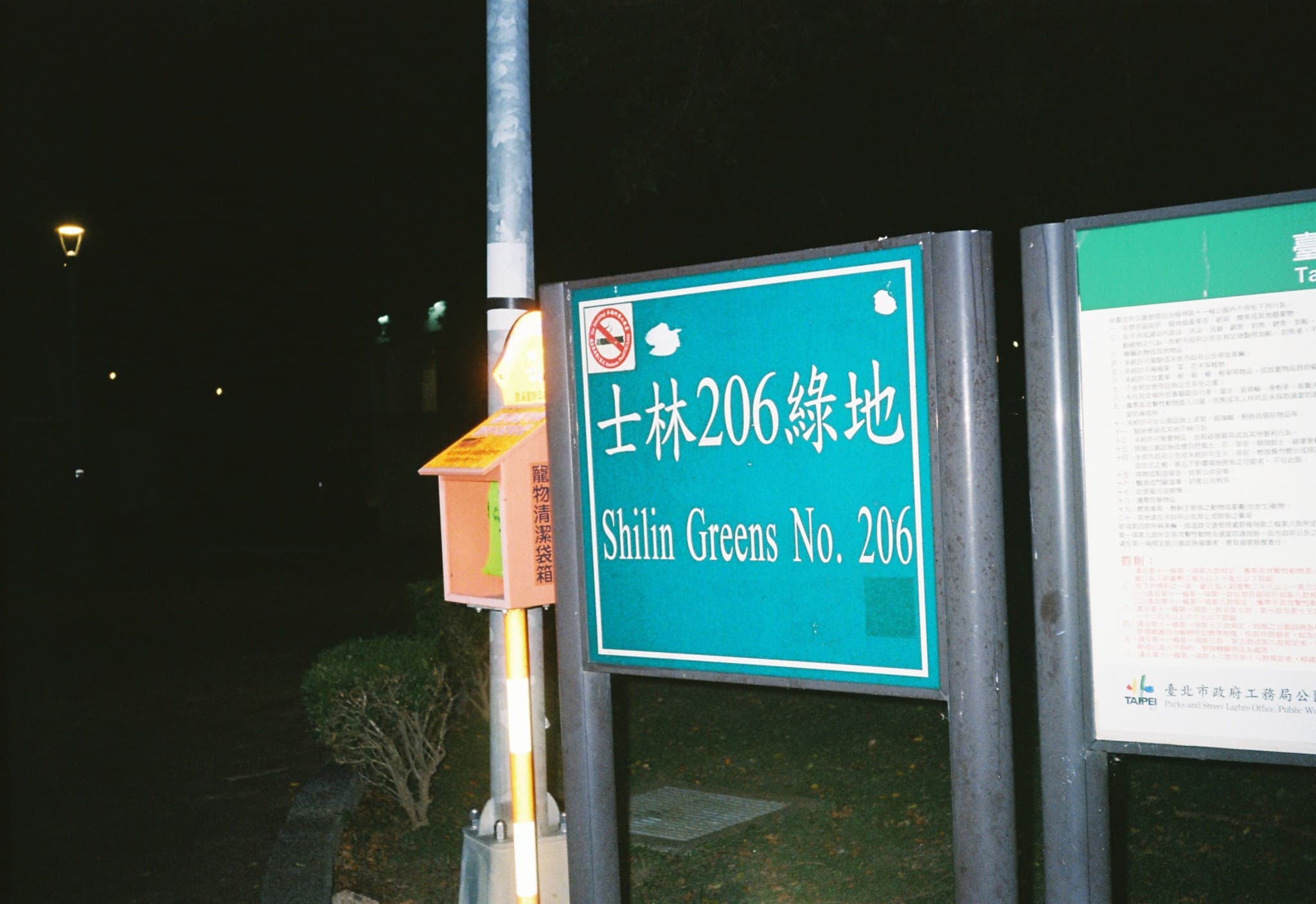 A teal sign labeled "Shilin Greens No. 206" stands on a post at night next to a pet waste bag dispenser.