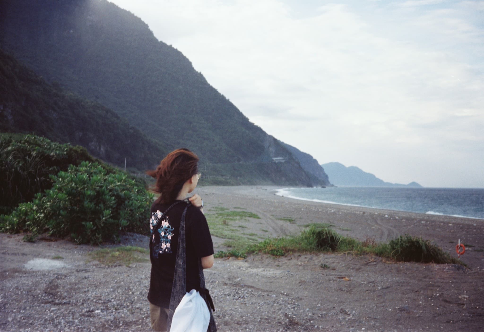 A person wearing a black floral shirt stands on a gravelly beach facing the ocean, with a large green mountain to their left.