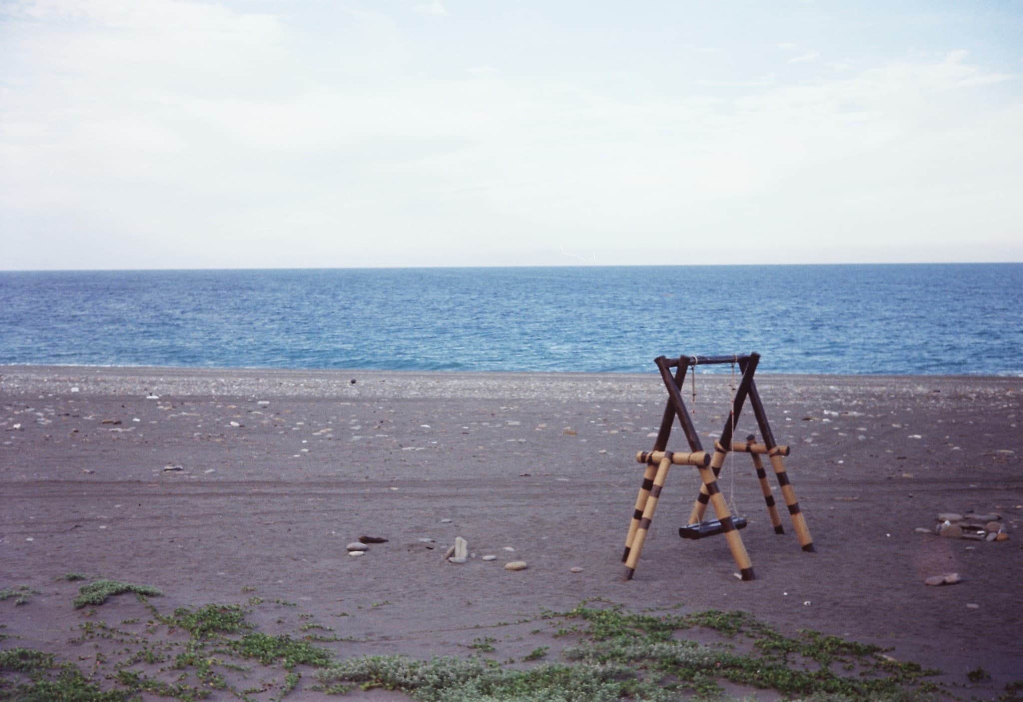 An empty wooden swing set stands alone on a sandy beach with a calm blue ocean in the background.