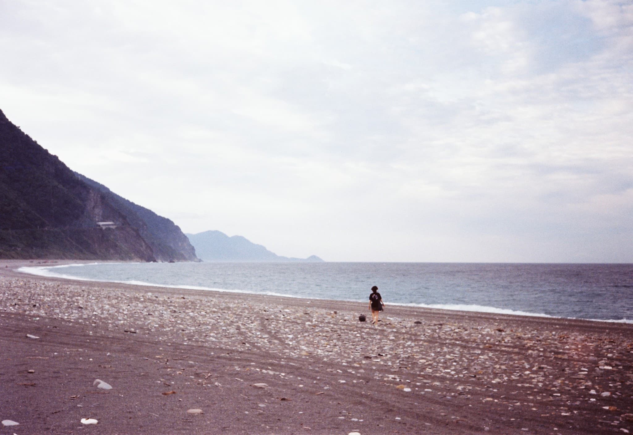A lone person walks along a wide, pebbly beach next to a calm ocean, with a steep, tree-covered cliff rising on the left side.