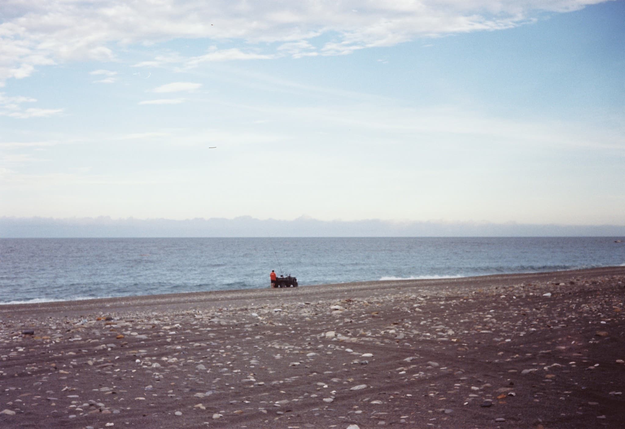 A person in an orange jacket stands next to a vehicle on a pebbly beach near the water's edge, with a calm sea and distant mountains in the background.