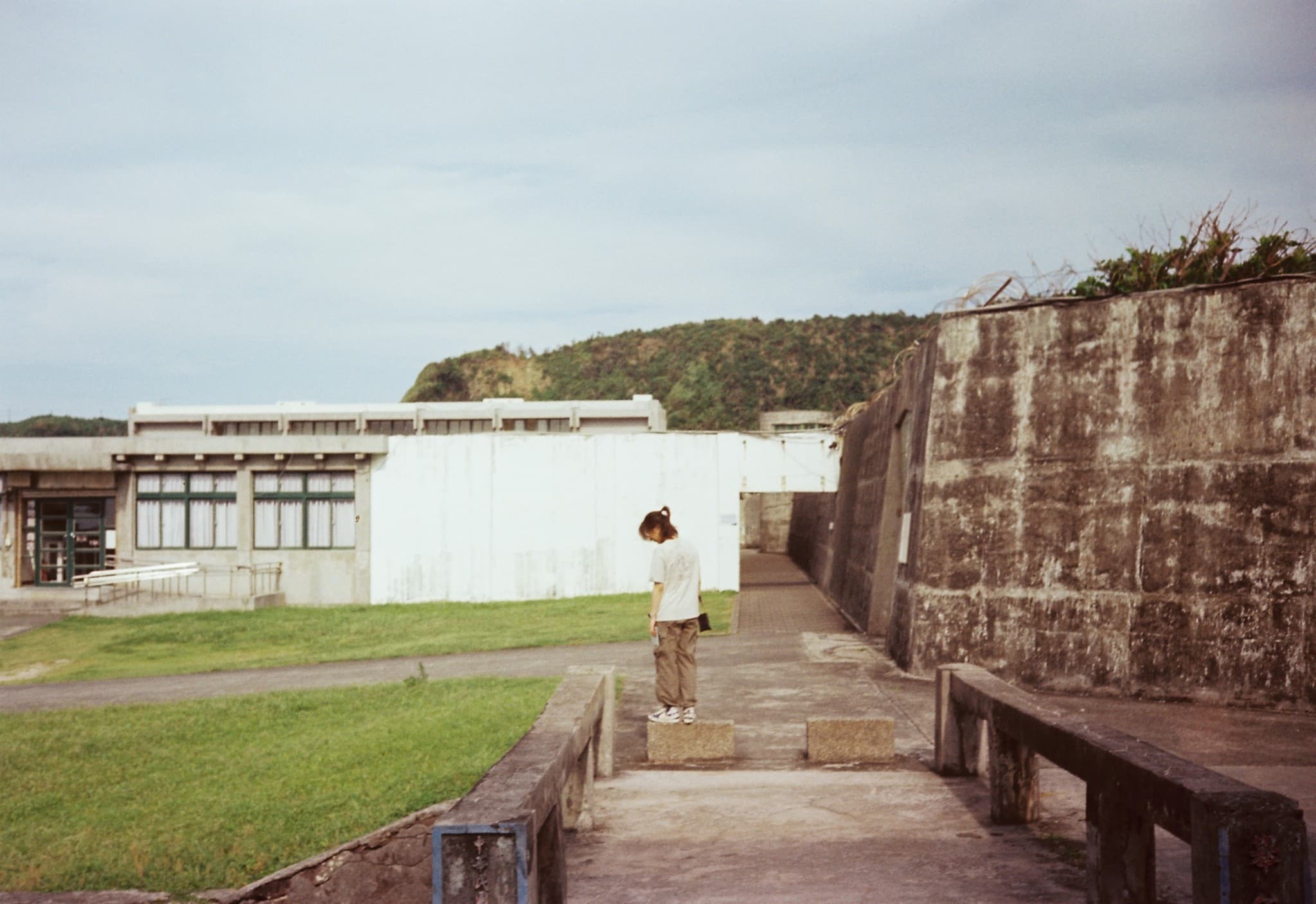 A person stands on a concrete walkway outdoors, positioned between a white building with windows on the left and a tall, weathered concrete wall on the right.