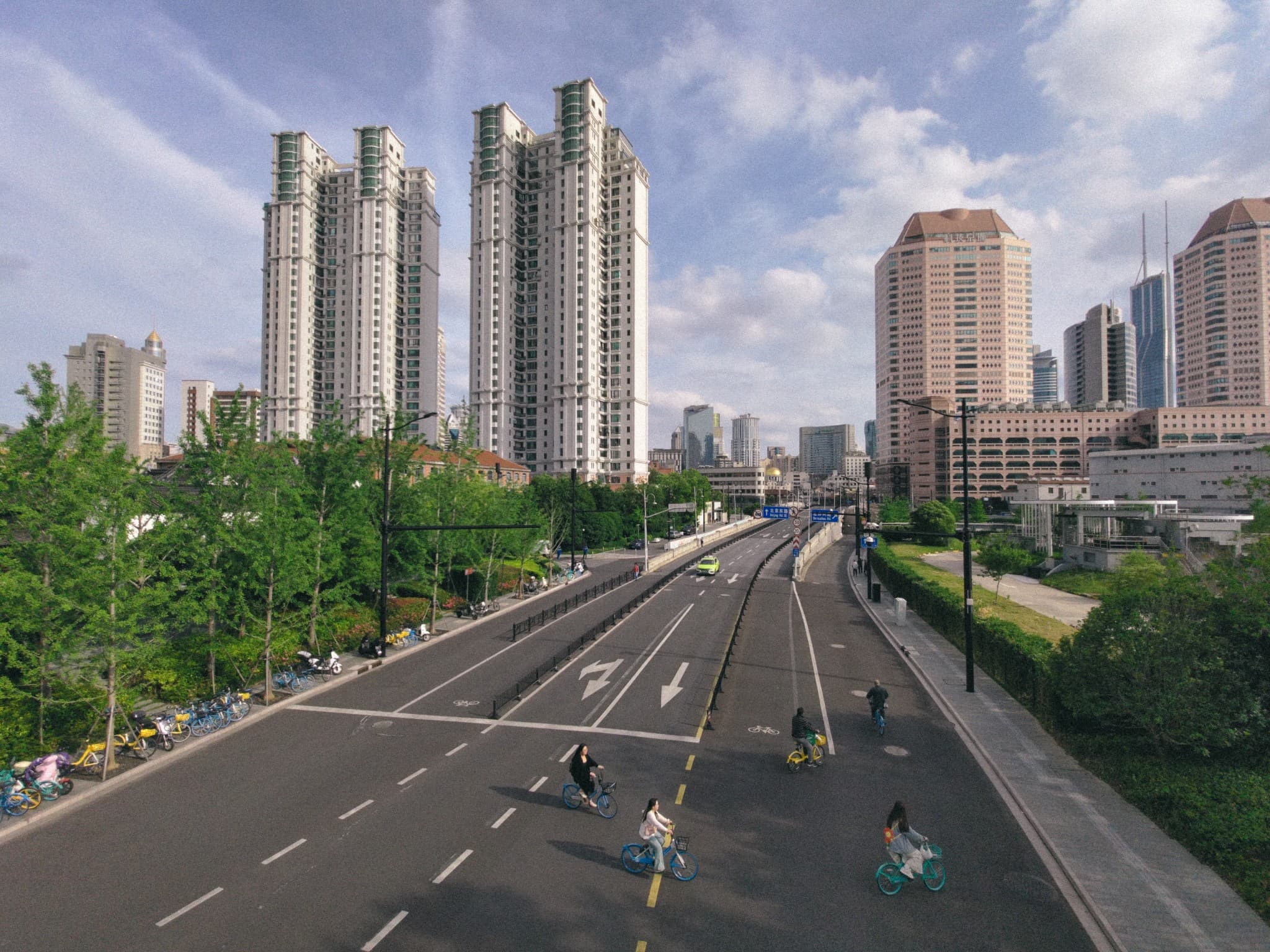 A wide urban street features several cyclists riding on a dedicated lane, surrounded by lush green trees and flanked by tall high-rise residential buildings under a partly cloudy blue sky.