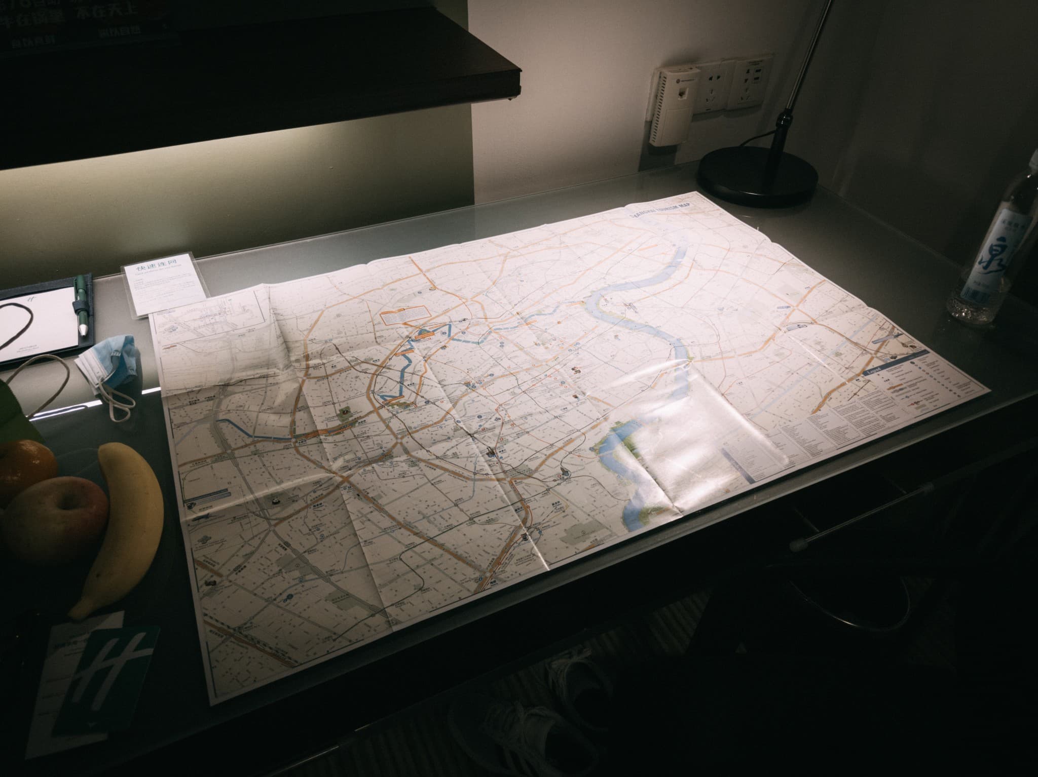 An unfolded paper map is spread across a dark desk under a lamp, surrounded by a banana, an apple, a face mask, and a water bottle.