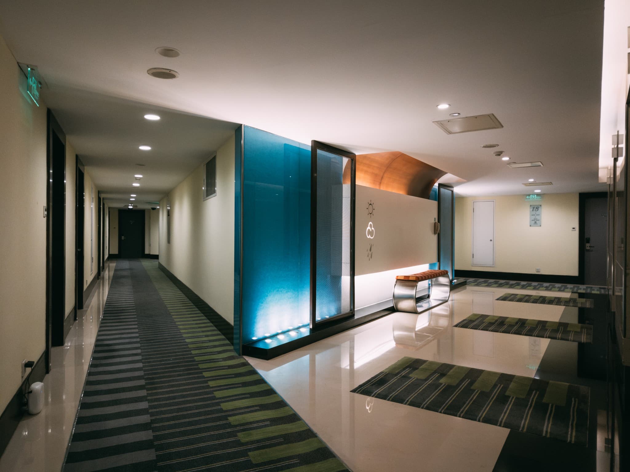 A long, modern hallway features a bright blue glass partition wall and a bench, with doors lining the left side and patterned carpets on the floor.