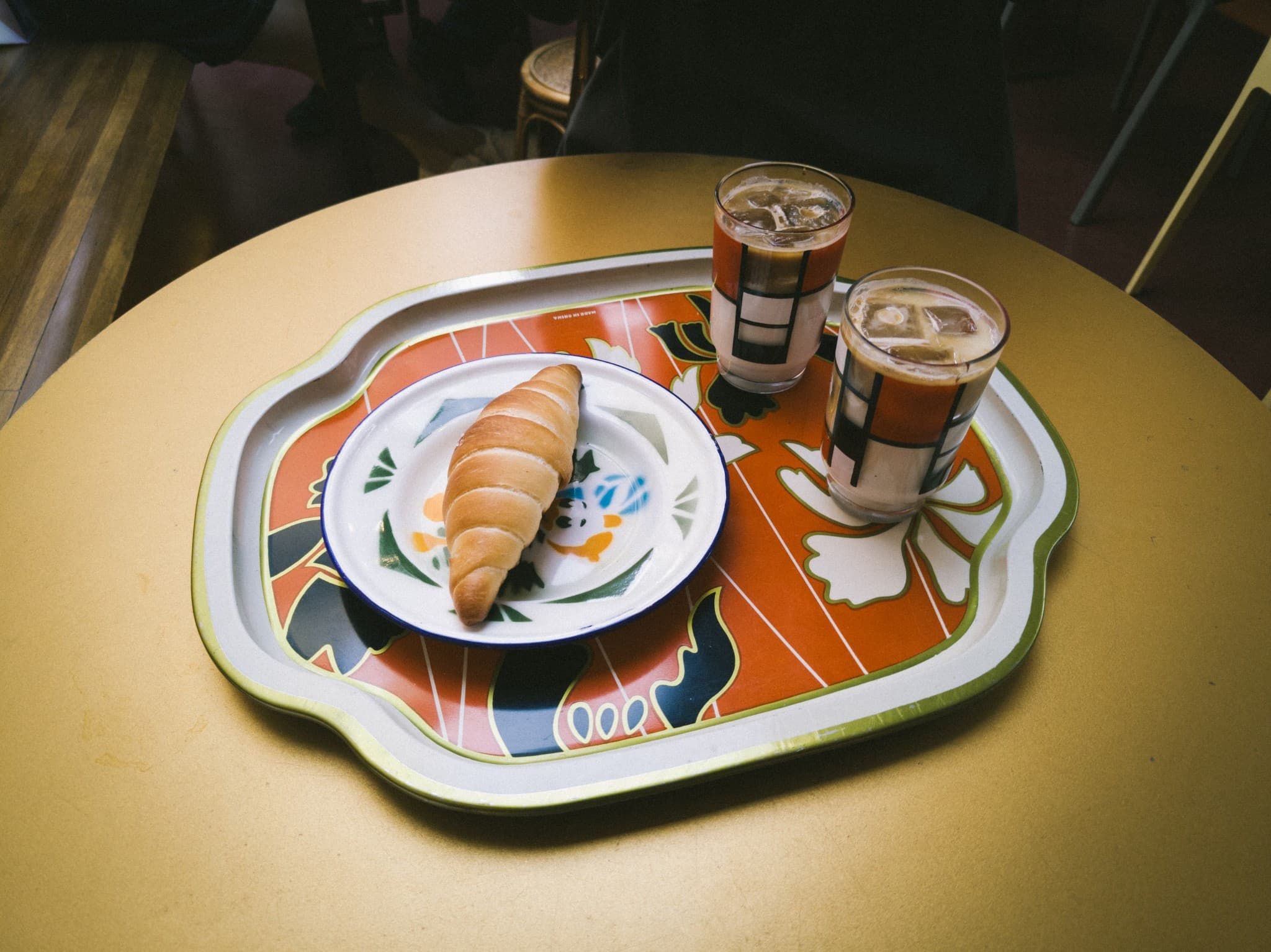 A colorful patterned tray holds a small plate with a croissant and two glasses of iced coffee.