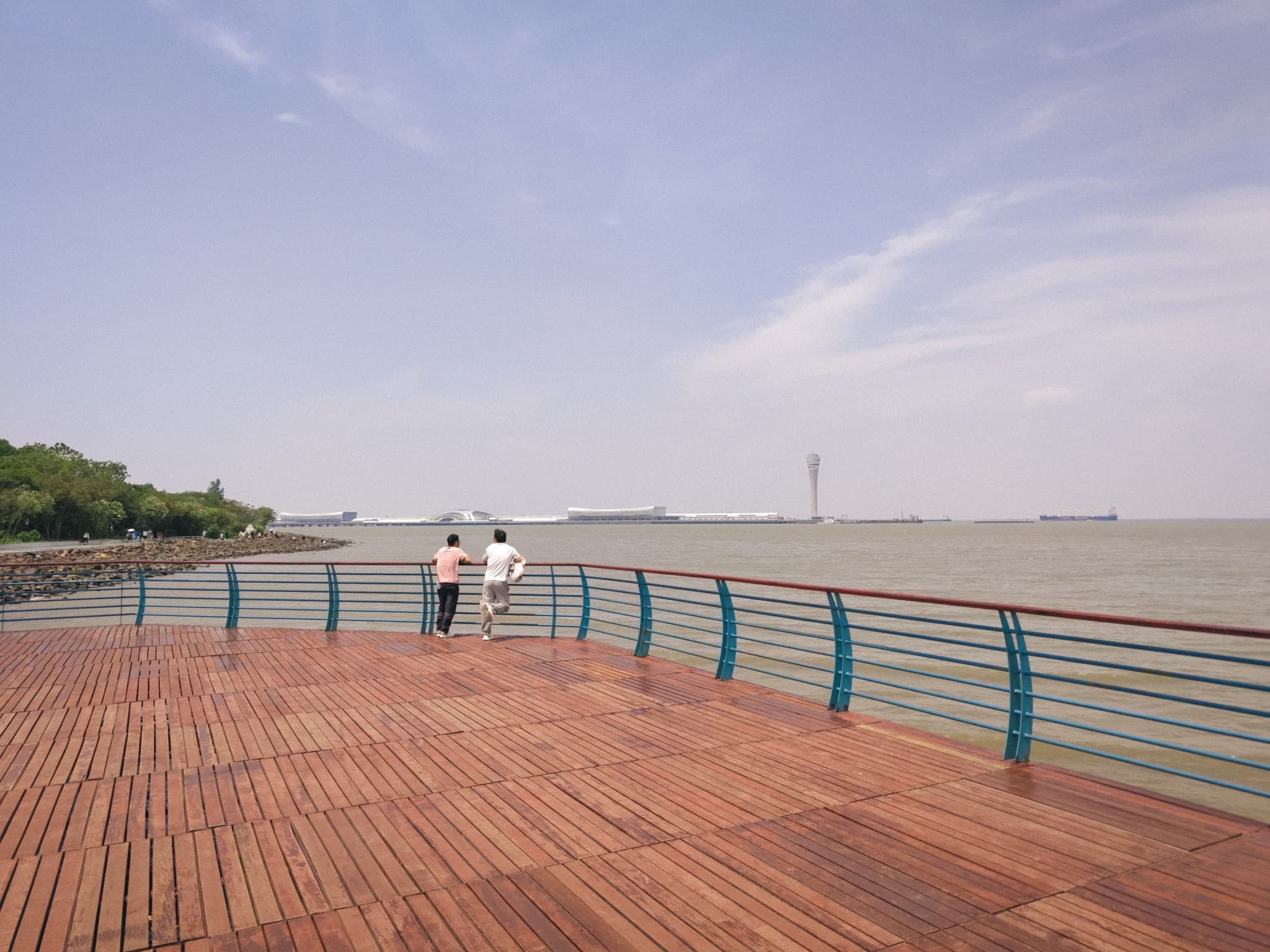 Two people stand on a wooden deck looking out over a large body of water toward a distant airport terminal and control tower under a blue sky.