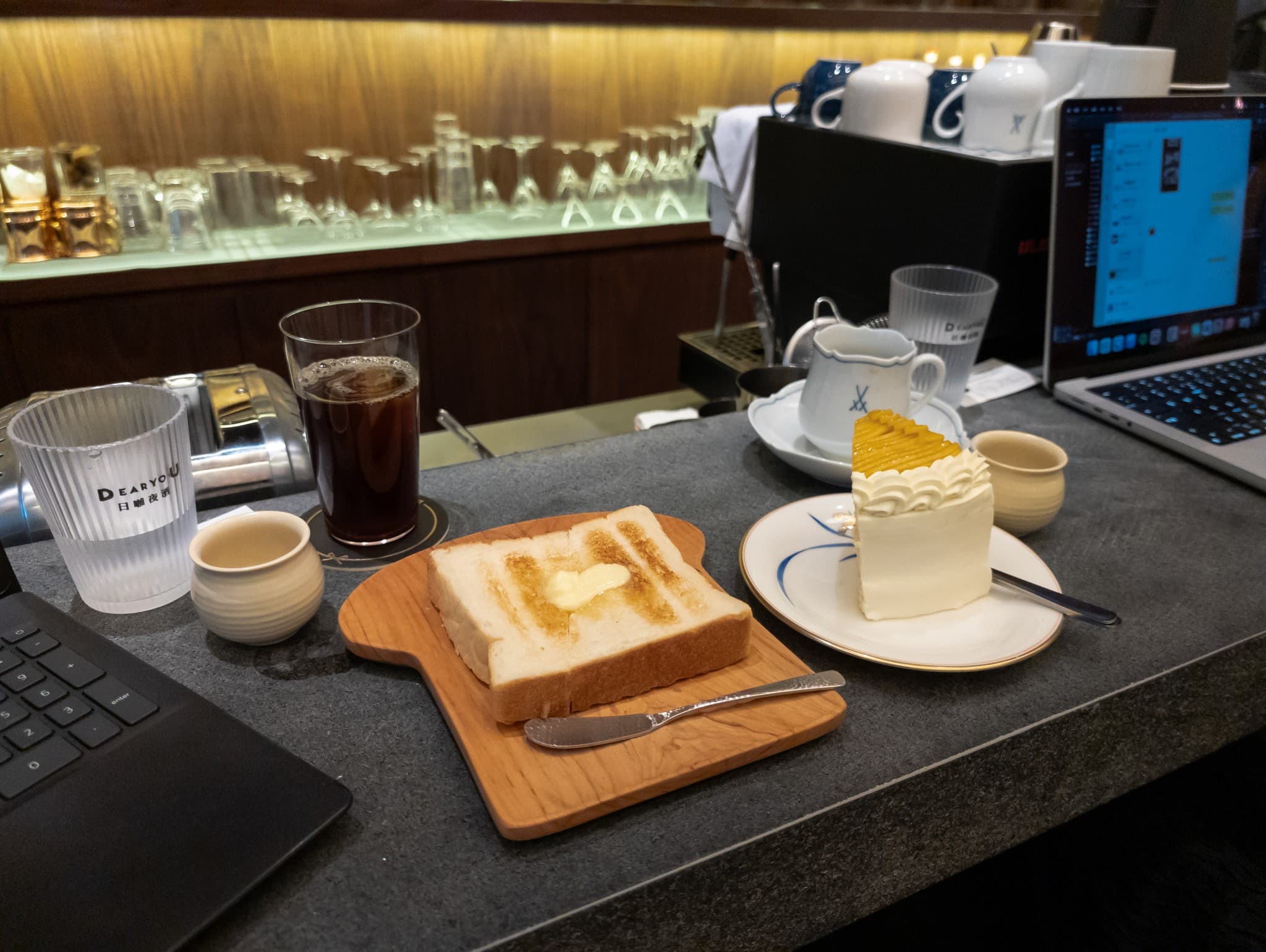 A slice of buttered toast on a wooden board sits next to a piece of white cake and a glass of iced coffee on a cafe counter with two laptops.
