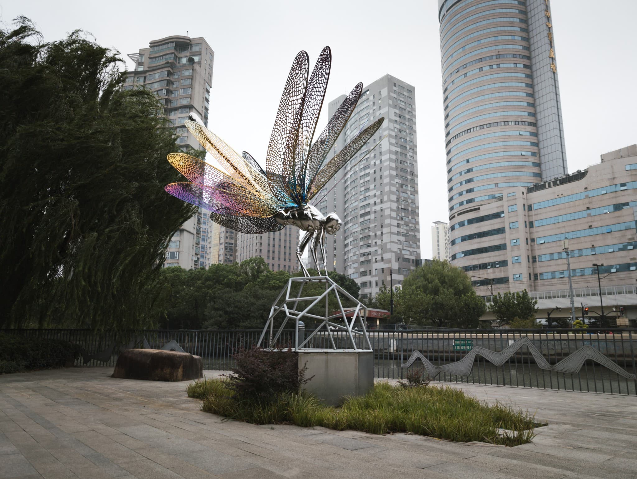 A large, metallic dragonfly sculpture with iridescent, colorful mesh wings stands on a geometric pedestal in an urban park with tall buildings in the background.