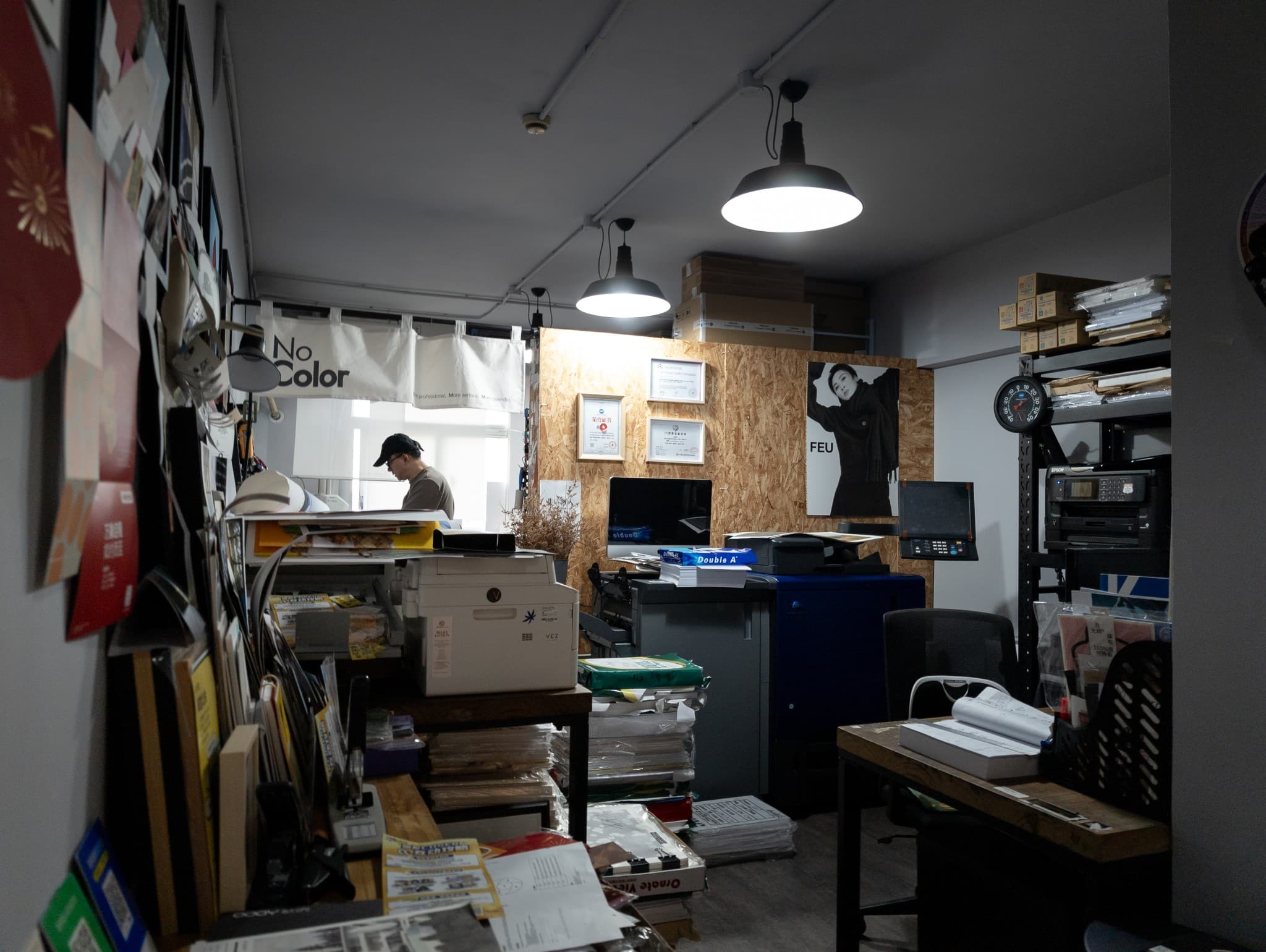 This image shows a cluttered office or print shop workspace featuring industrial pendant lights, stacks of paper and boxes, printers, a wooden wall with framed certificates, and a person working in the background near a window.