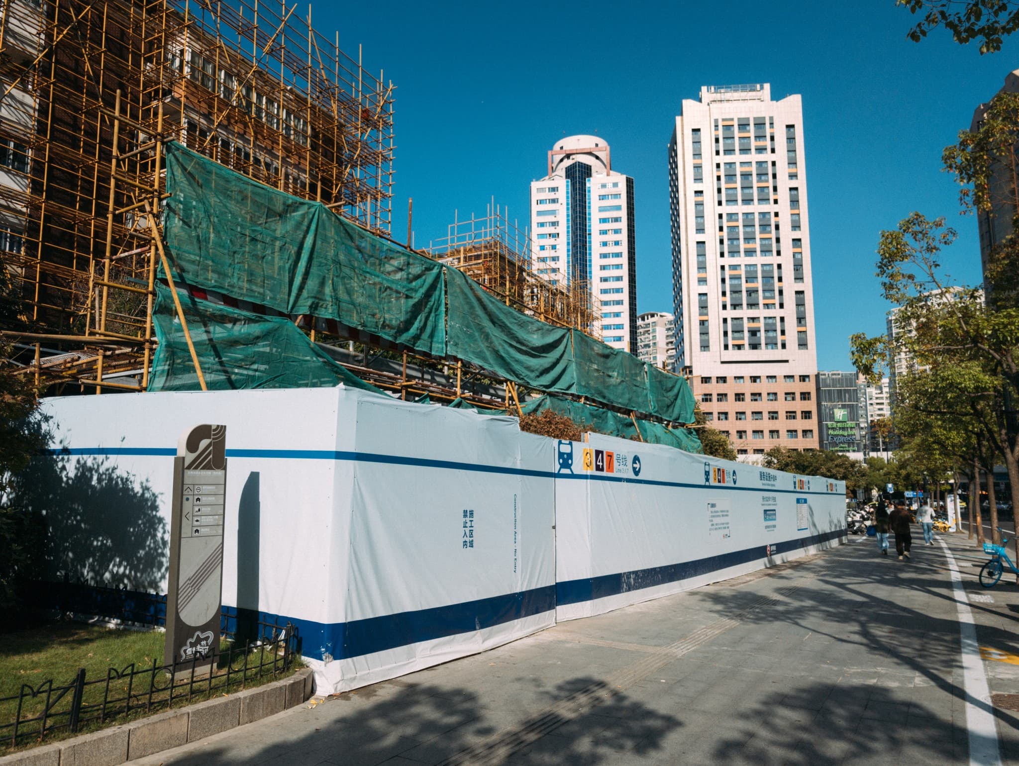 A white construction barrier with blue stripes and subway line icons stands in front of scaffolding covered in green safety netting, with tall white skyscrapers visible in the background under a clear blue sky.