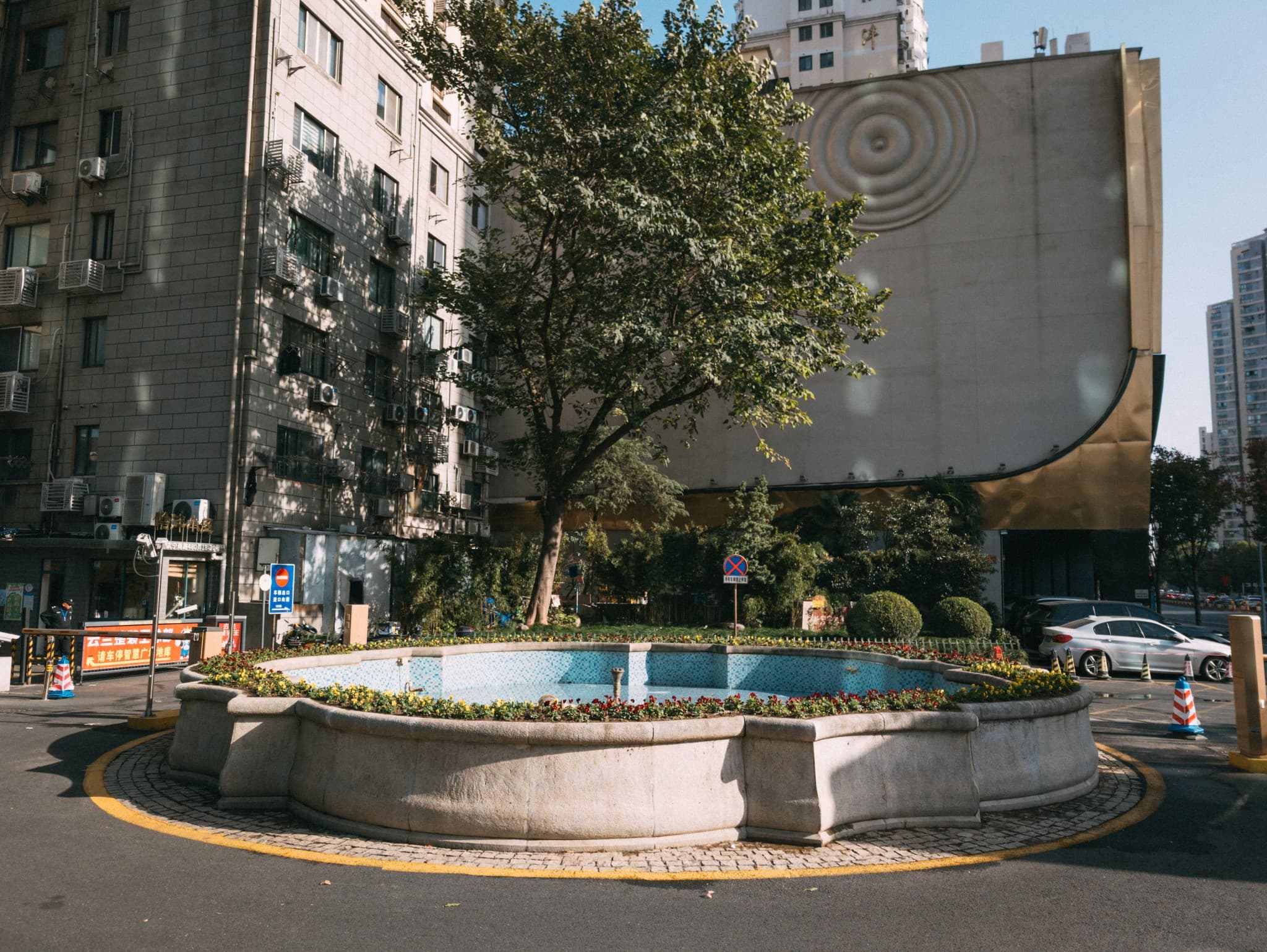 A large, stone fountain surrounded by flower beds sits in a paved area, framed by a leafy tree and tall urban buildings in the background.
