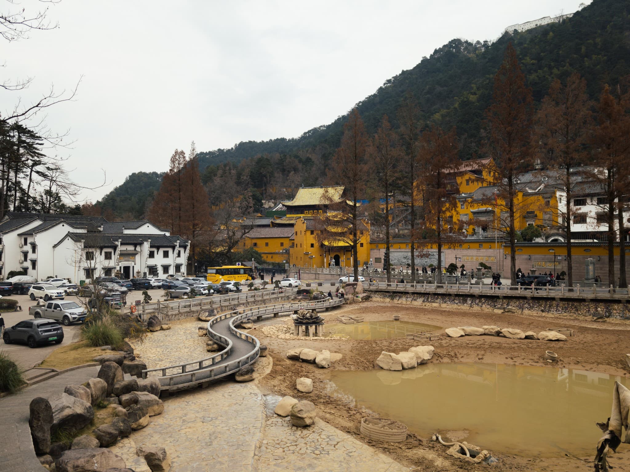 A scenic view of a traditional yellow temple complex nestled at the foot of a forested mountain, featuring a dry pond with a winding walkway in the foreground and white buildings to the left.