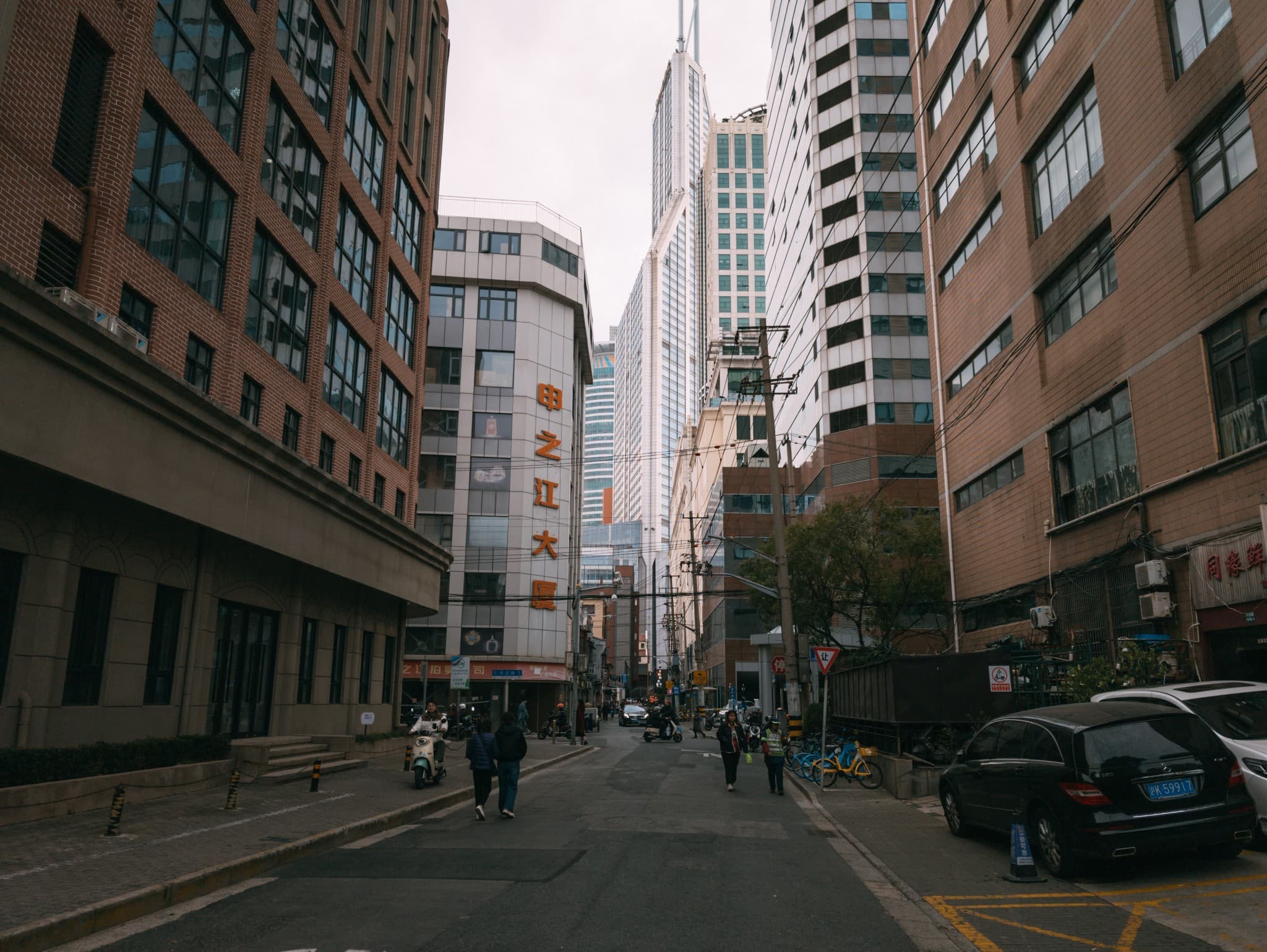 Pedestrians walk and ride scooters along a city street flanked by tall brick and modern buildings, with a black car parked on the right side.