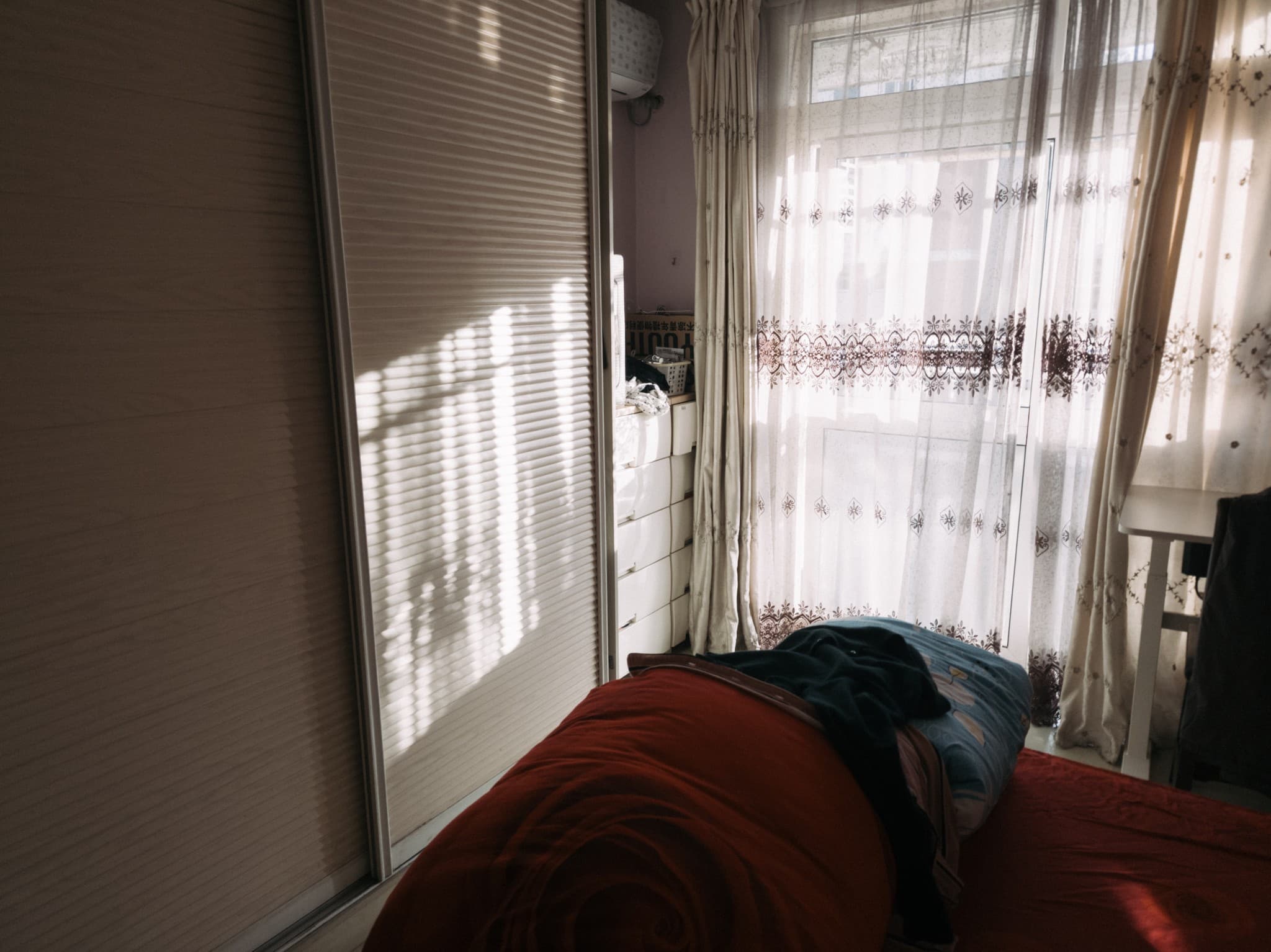 This image shows a bedroom interior with a bed covered in red sheets in the foreground, a white wardrobe with sliding blinds on the left, and a window with sheer curtains letting in natural light on the right.
