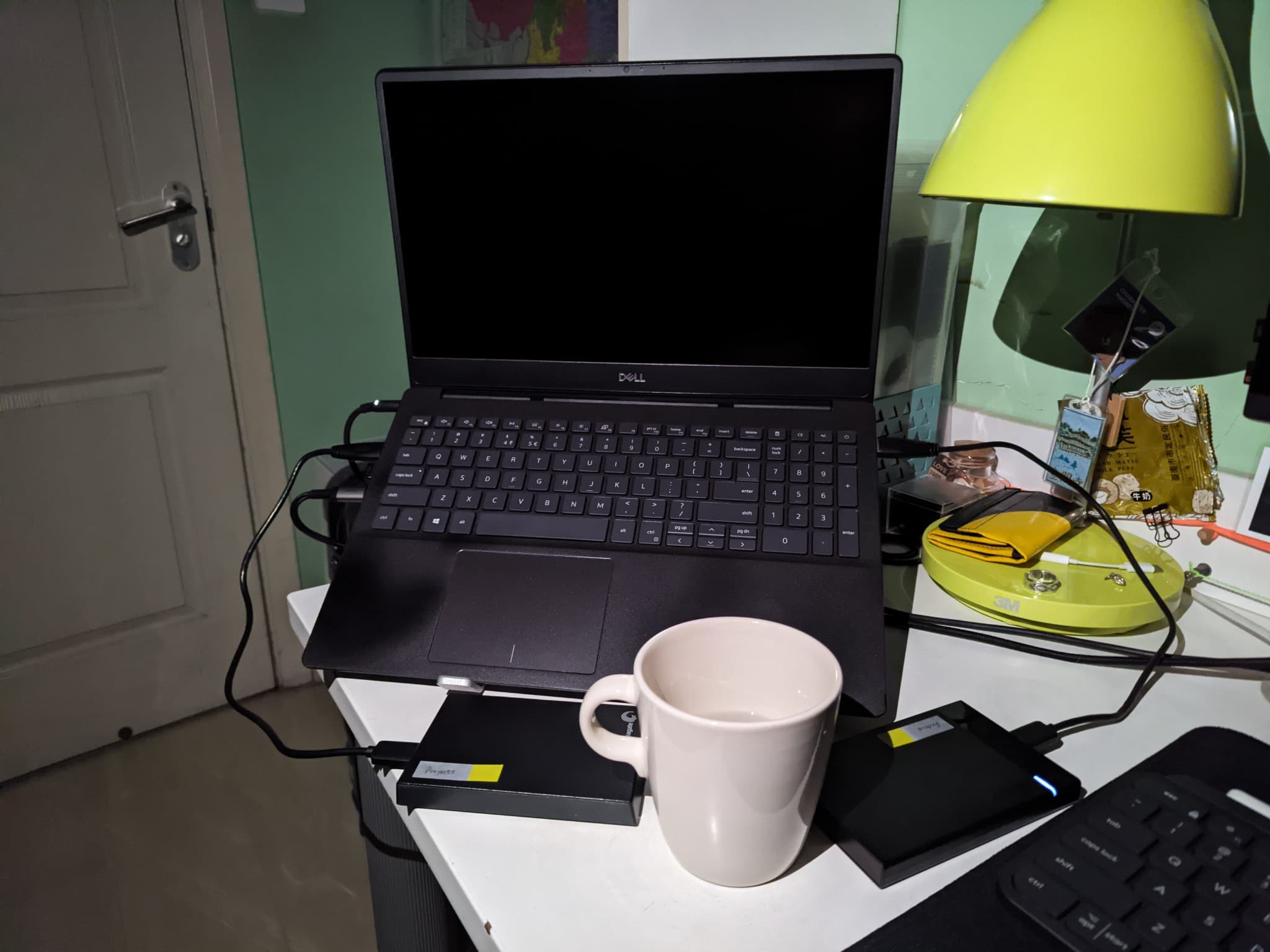 A black Dell laptop sits open on a white desk next to a white mug and external hard drives, illuminated by a bright yellow lamp.