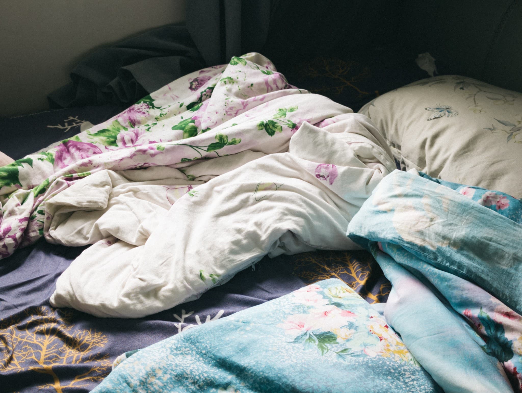 An unmade bed featuring a rumpled white duvet, pink floral sheets, and blue blankets resting on a dark blue sheet with a gold tree pattern.