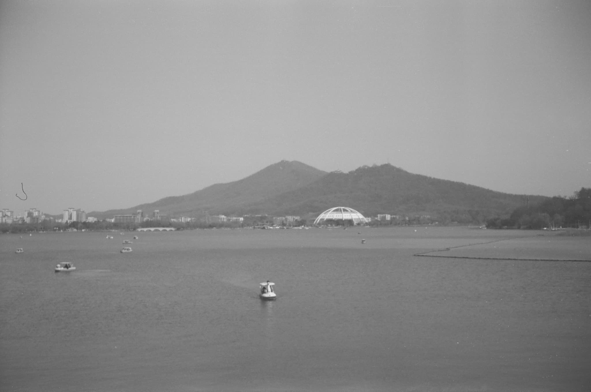 This black and white photograph shows a large lake with several small boats on the water, backed by a city skyline, a white domed structure, and two hills in the distance.