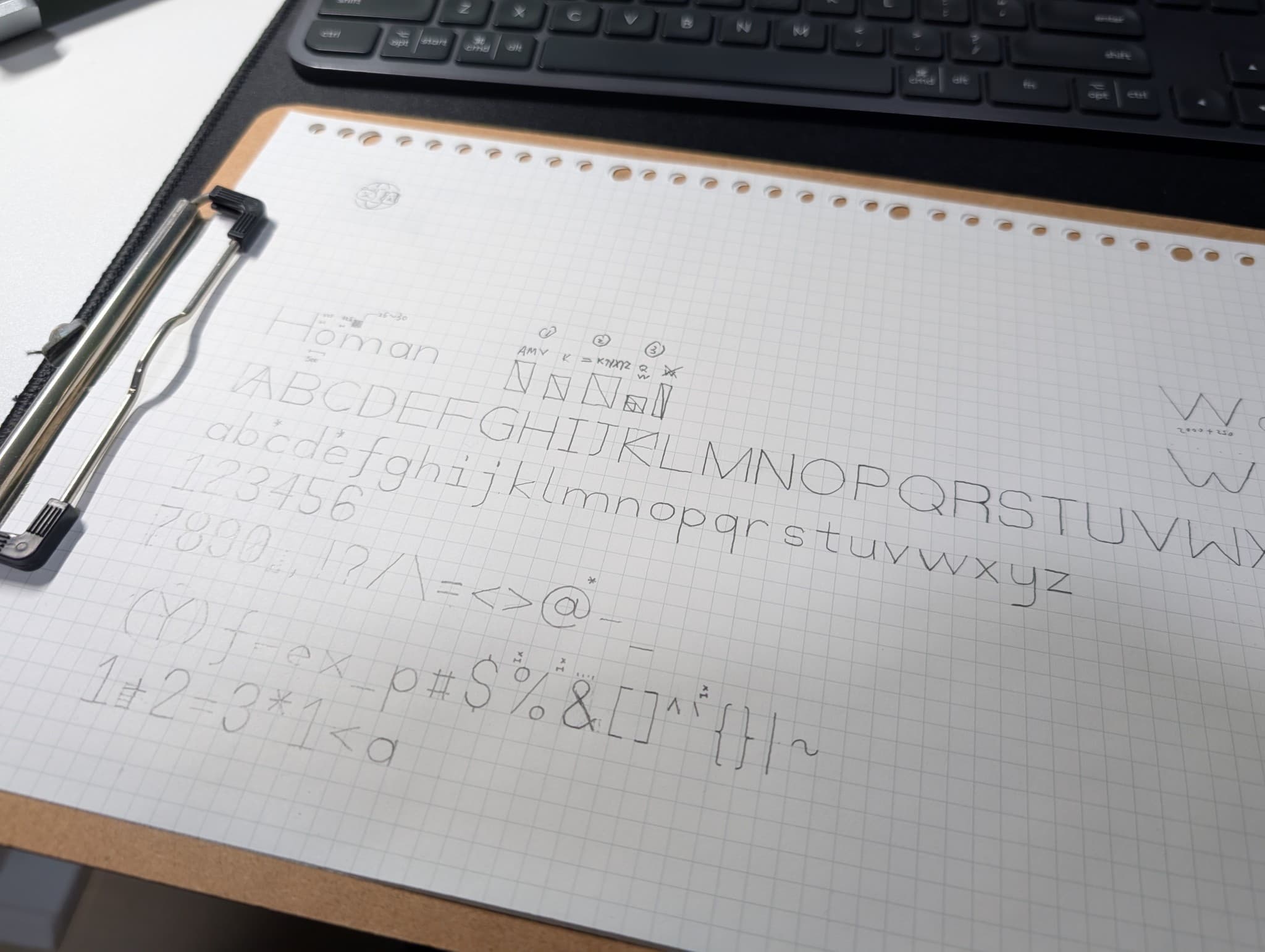 An overhead shot of a graph notebook page clipped to a clipboard, displaying a hand-lettered custom font alphabet including letters, numbers, and symbols, with a black keyboard visible at the top.